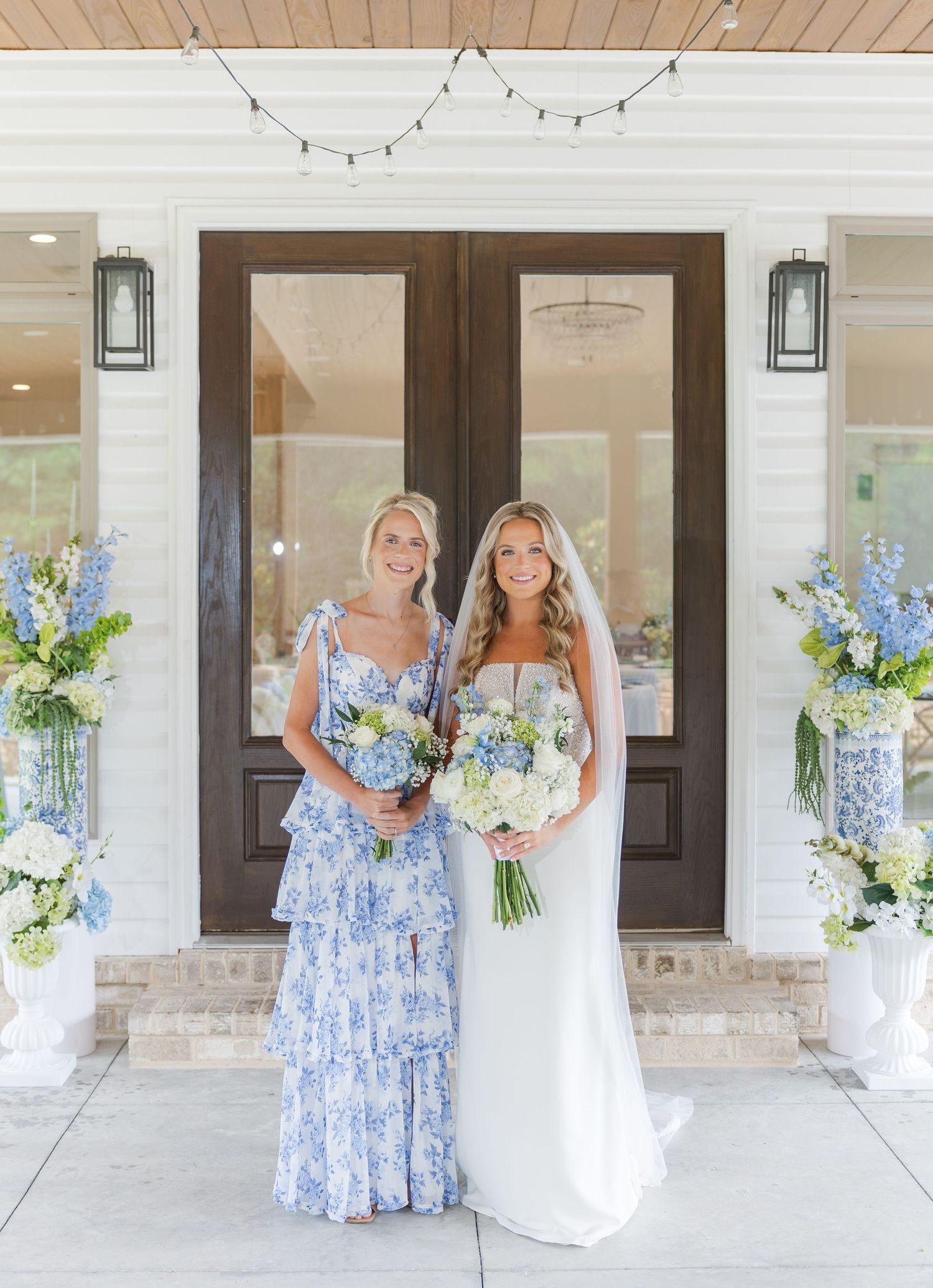 Bride in strapless white gown and woman in blue floral dress posing together in front of wedding venue doors.