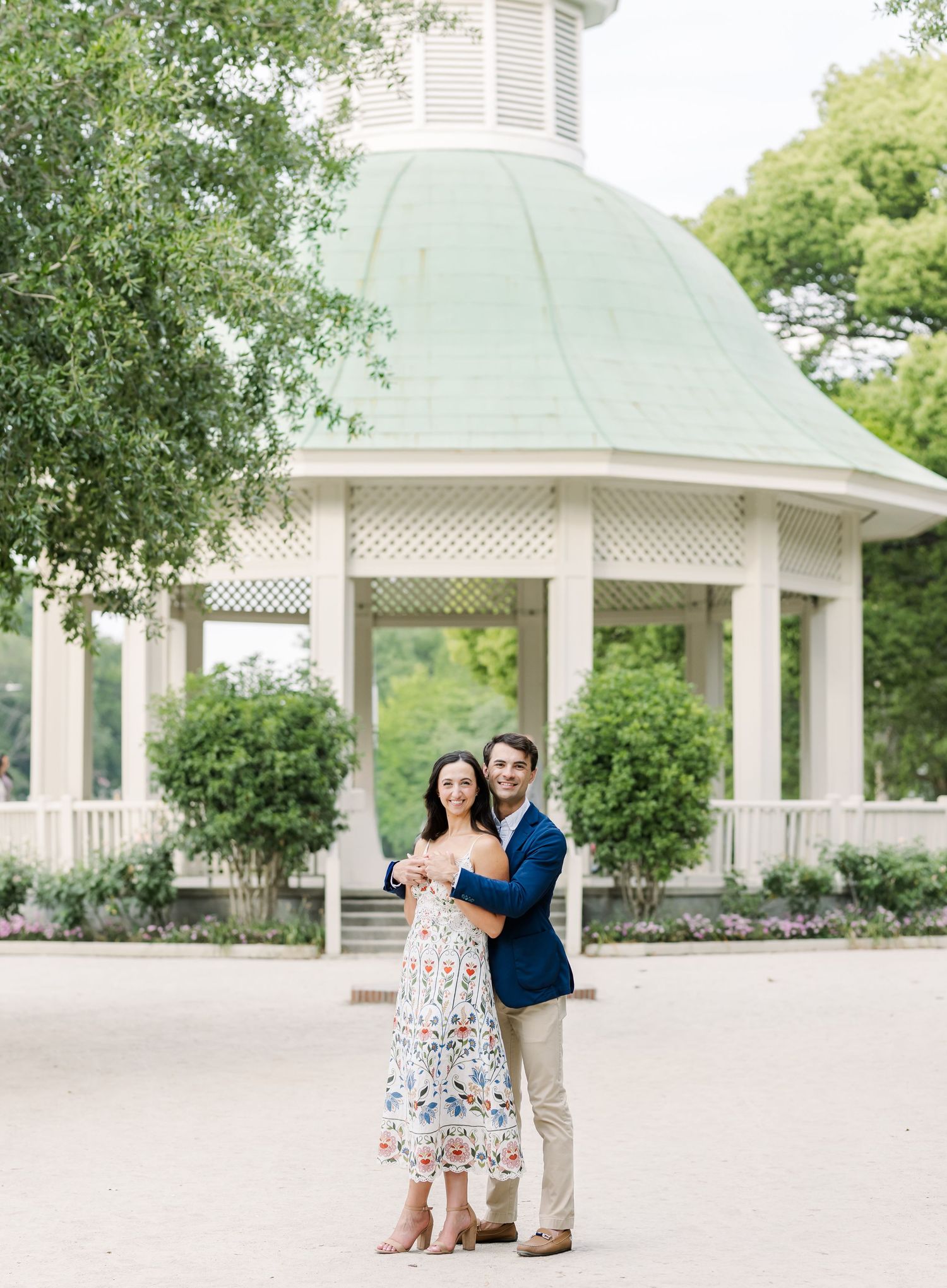 Couple embracing and smiling in front of a white gazebo with green dome roof during engagement photos.