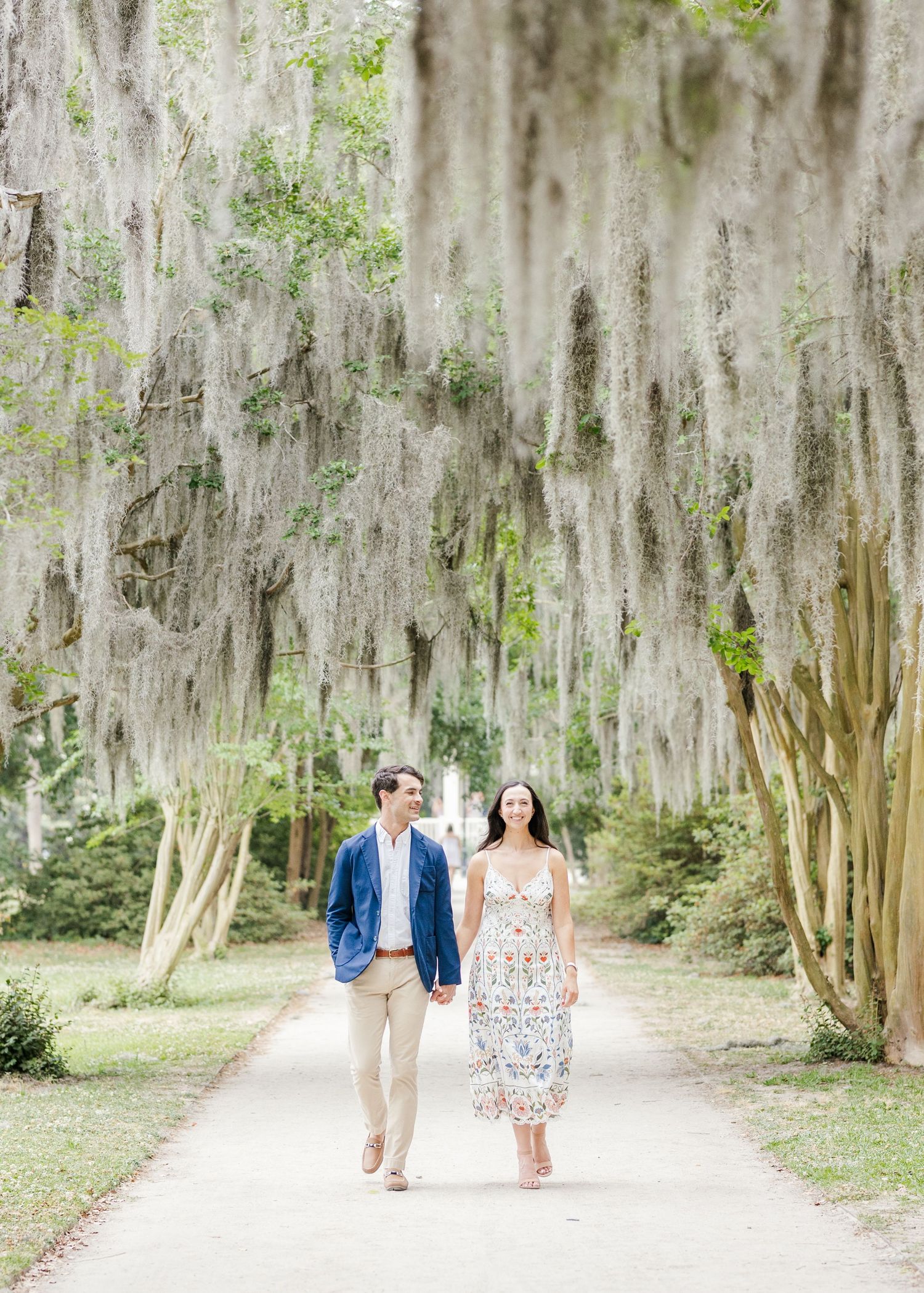 Couple walks hand-in-hand down a moss-draped oak tree path during an outdoor engagement session.