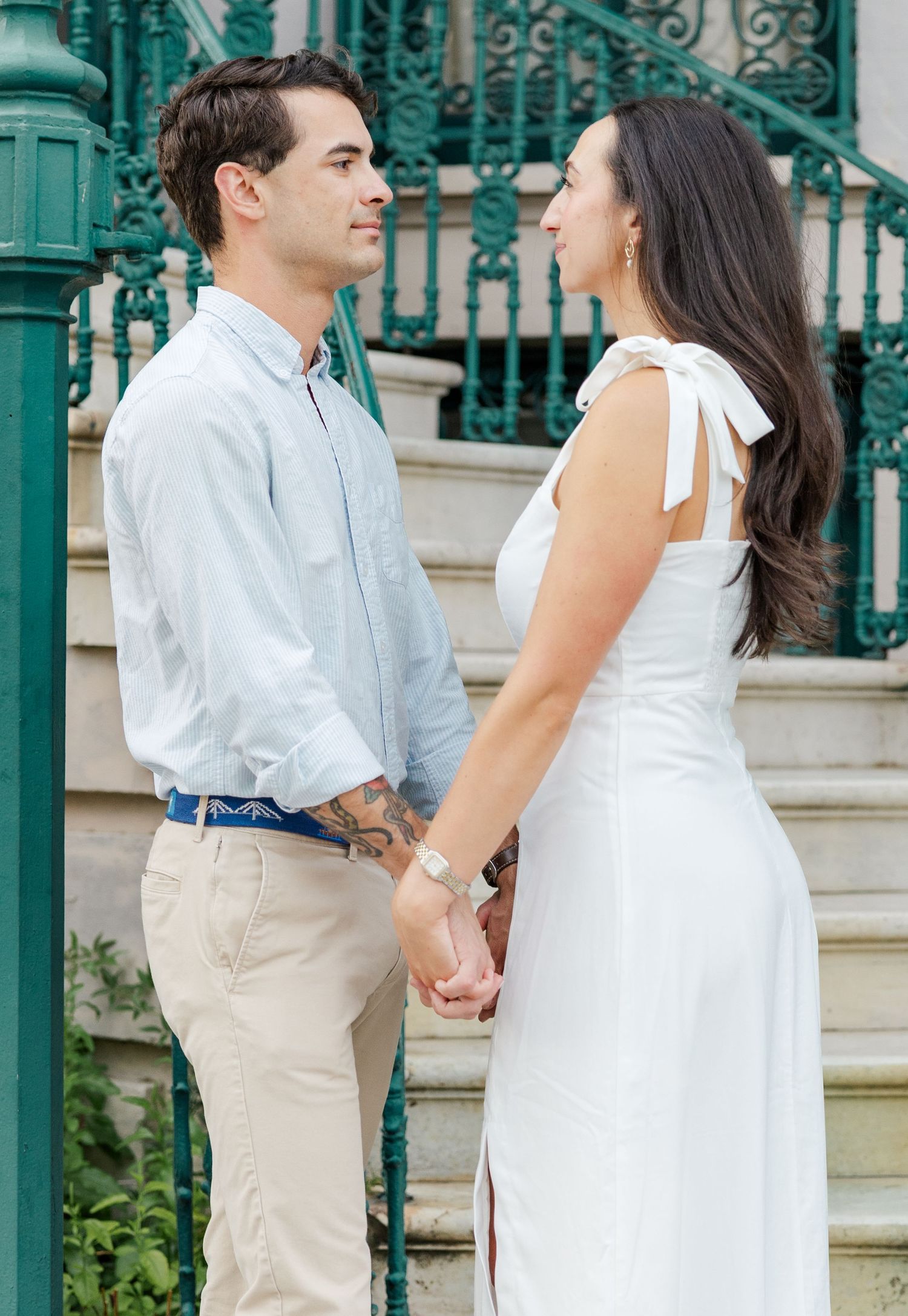 Engaged couple holding hands and gazing at each other near ornate teal iron railings and stone steps.