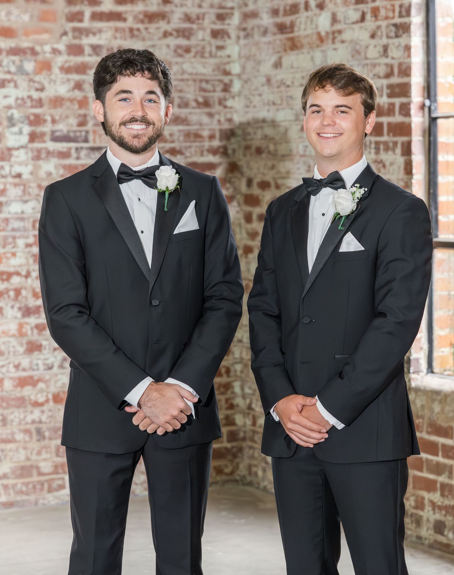 Two groomsmen in black tuxedos with white boutonnieres smiling before exposed brick wall at wedding.