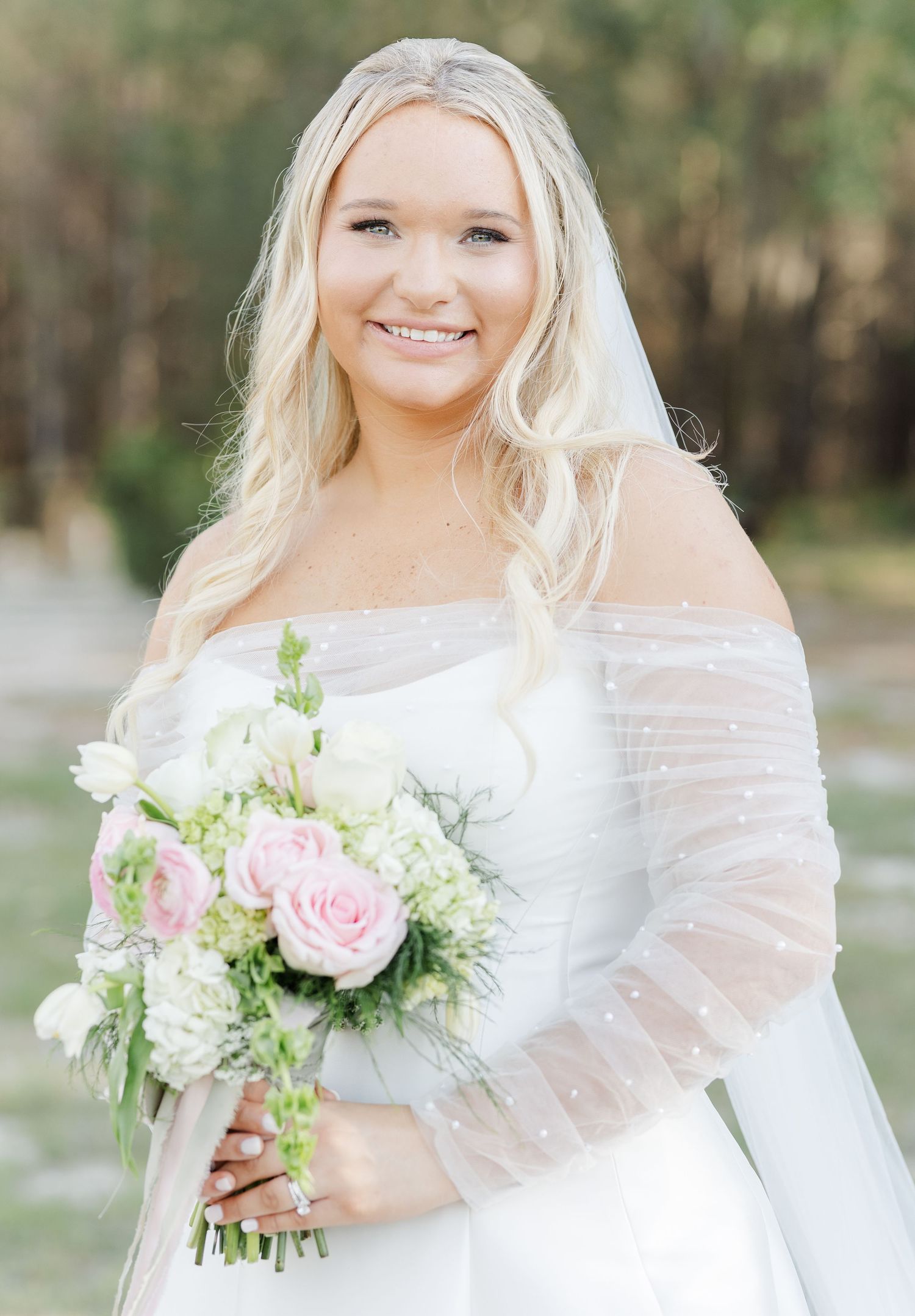 Smiling blonde bride in off-shoulder lace wedding gown holding pink rose and white hydrangea bouquet with veil.