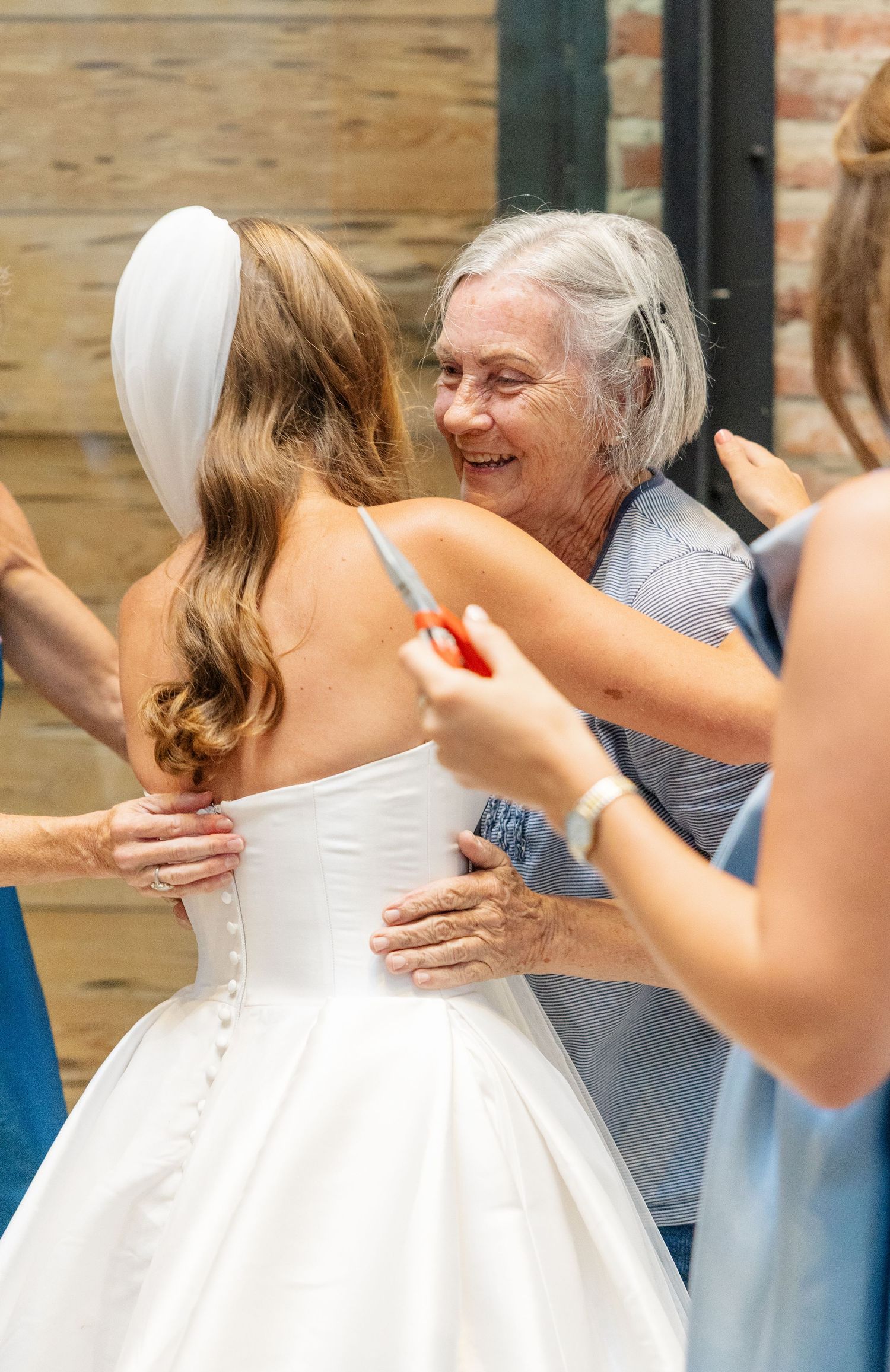 Older woman smiles while helping bride button up elegant white wedding gown with veil and long brown hair.