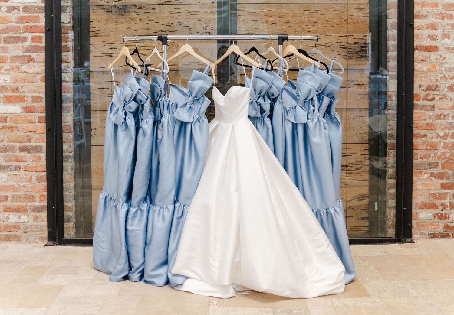 White wedding gown displayed beside dusty blue bridesmaid dresses hanging on rack near brick wall.
