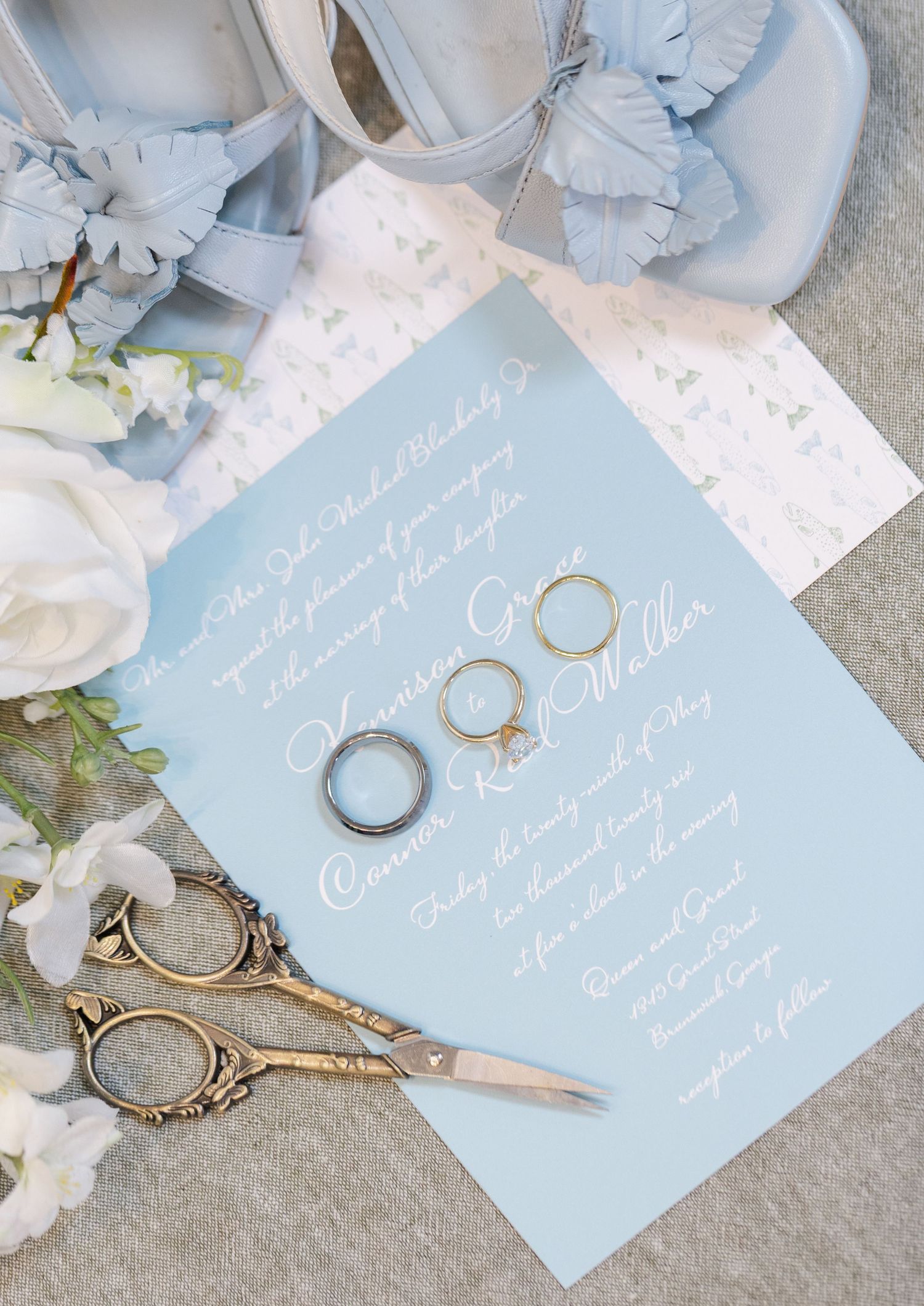 Light blue wedding invitation suite with gold rings, vintage scissors, white roses, and blue bow heels on a gray background.