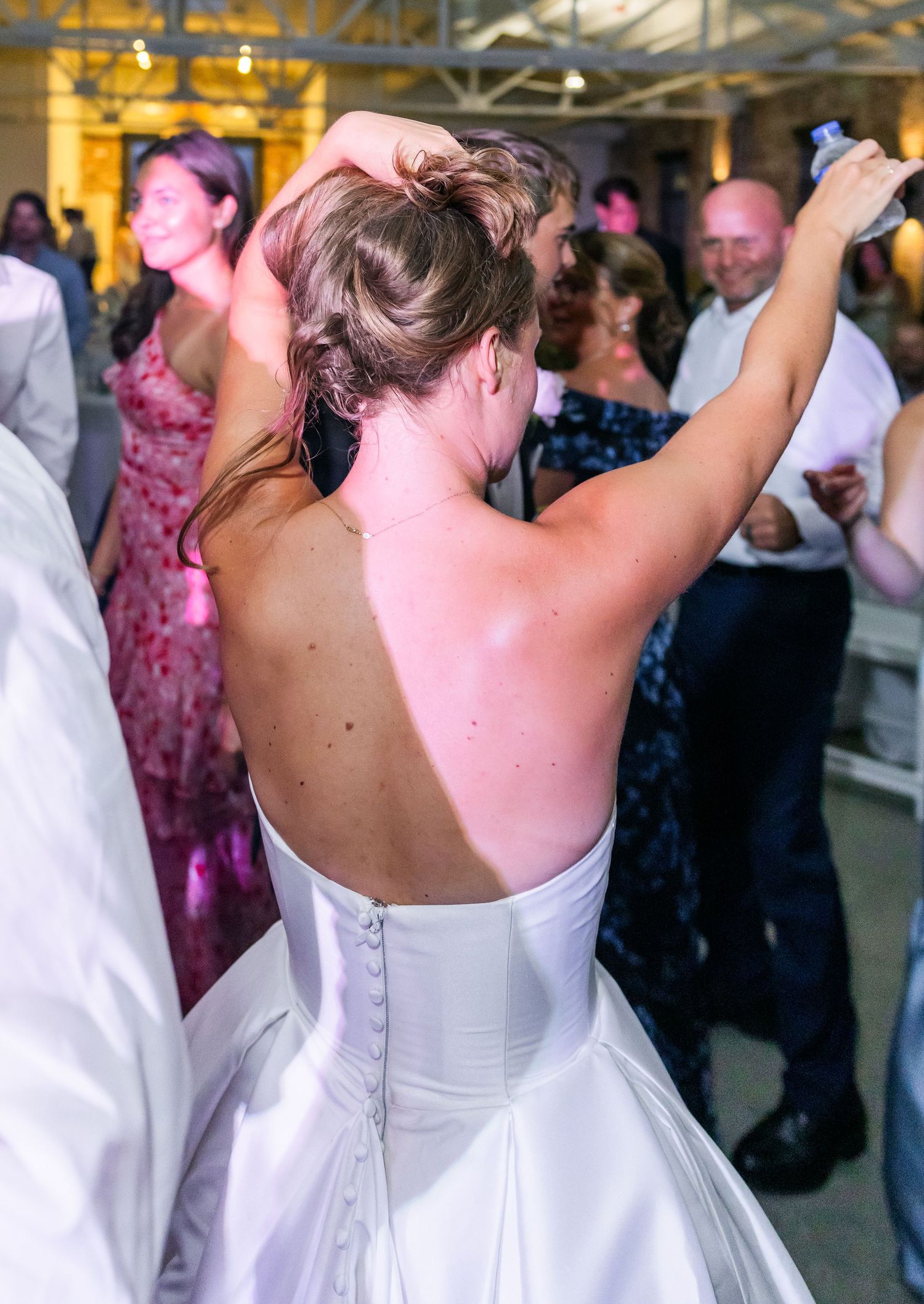 Bride in backless white gown dancing with guests at lively wedding reception.