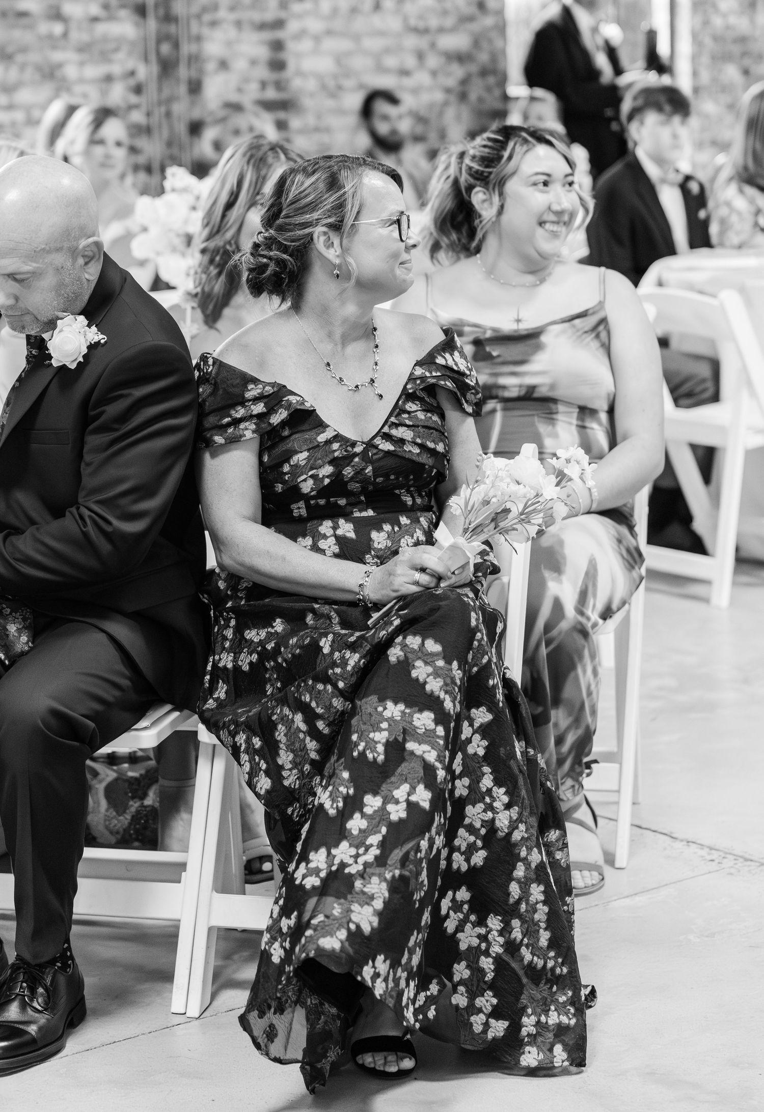 Black and white photo of smiling wedding guests seated in white chairs during an indoor ceremony.