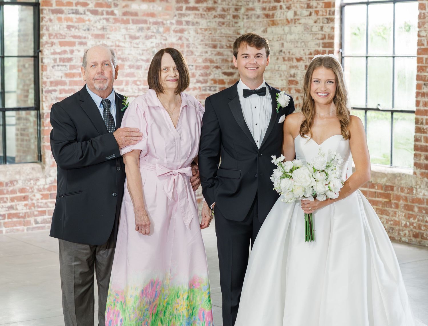 Wedding party of four poses together, bride in white gown with groom in tuxedo and two guests.