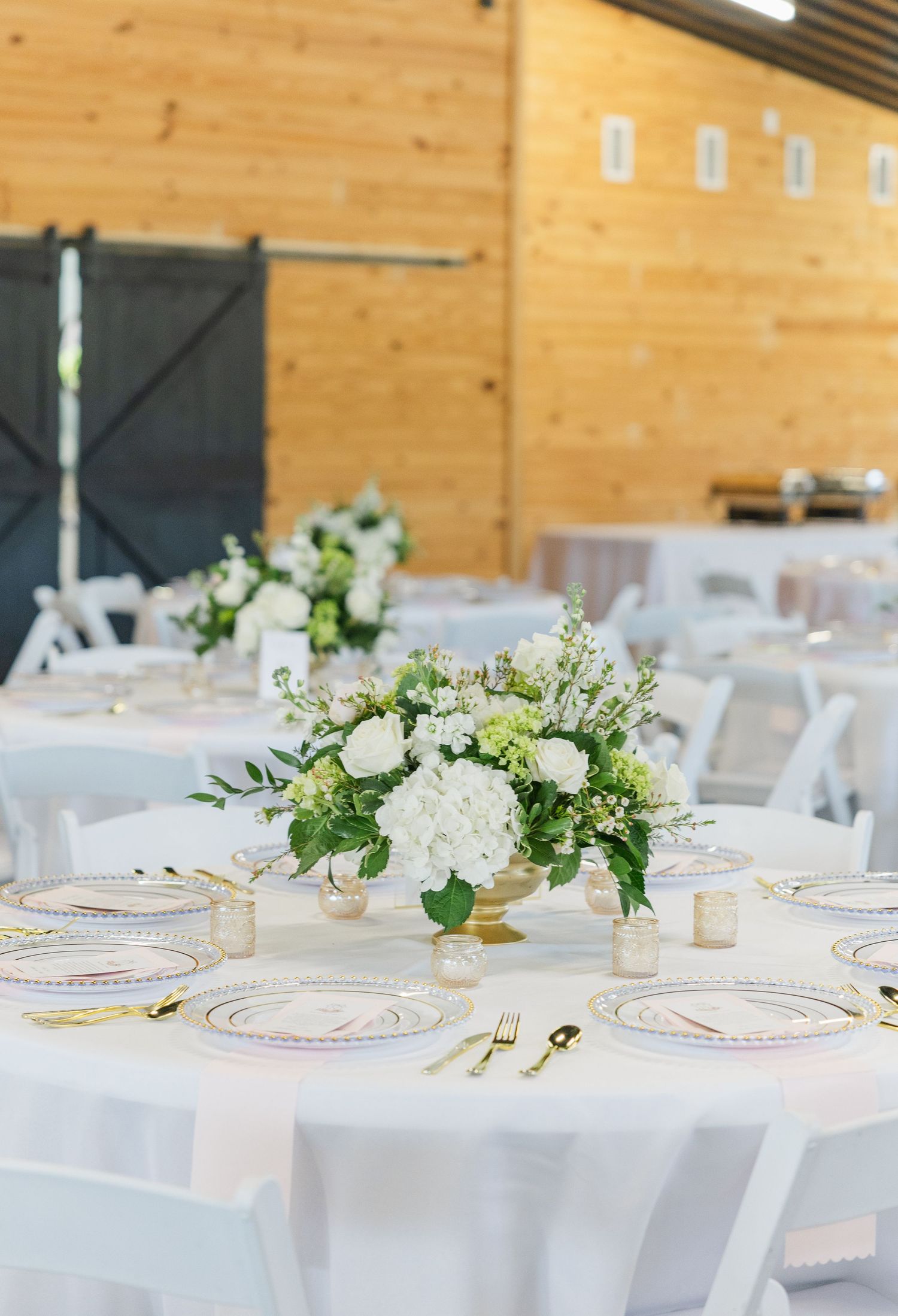 Elegant barn wedding reception with white round tables, gold centerpieces of white hydrangeas and greenery.