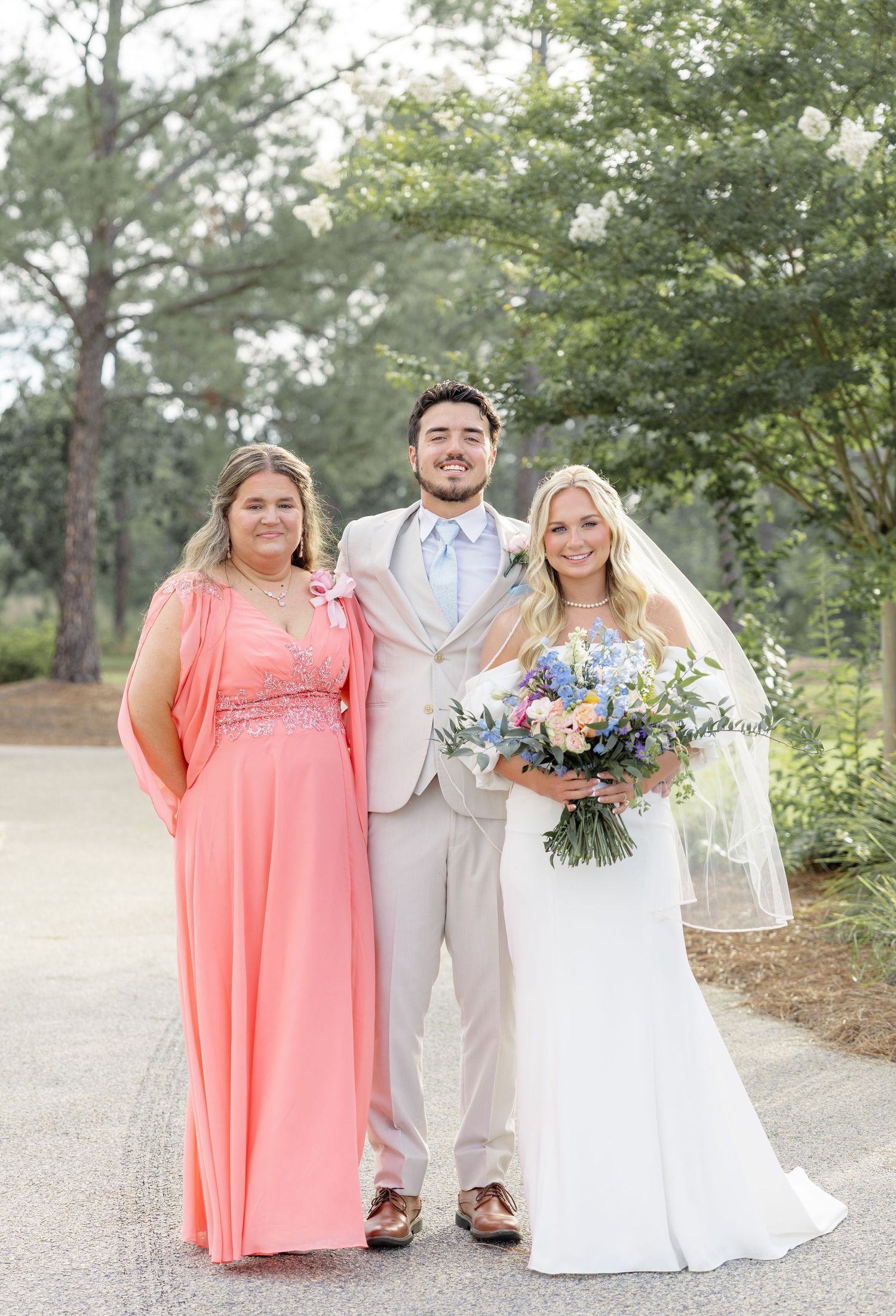 Bride in white gown with bouquet and groom in gray suit pose with woman in coral dress outdoors.