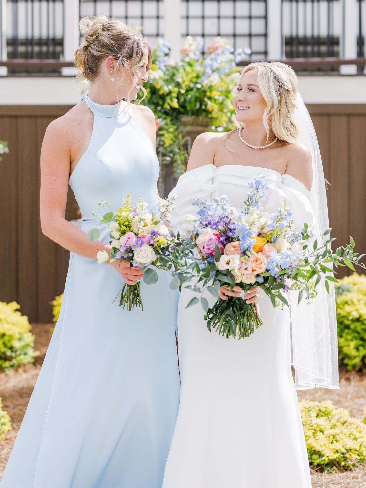 Bride in white strapless gown and bridesmaid in light blue halter dress holding colorful spring bouquets outdoors.