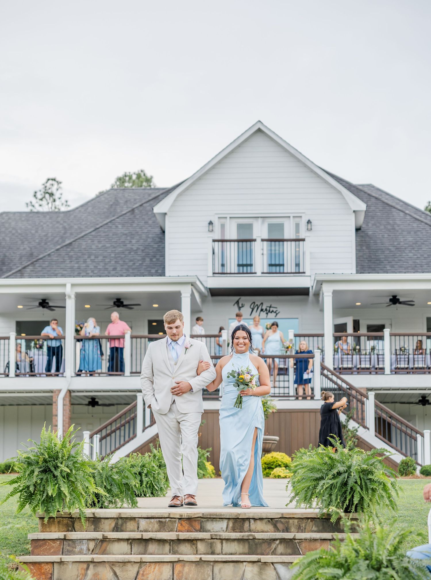 Groomsman in tan suit escorts bridesmaid in blue gown down steps at elegant white farmhouse wedding venue.