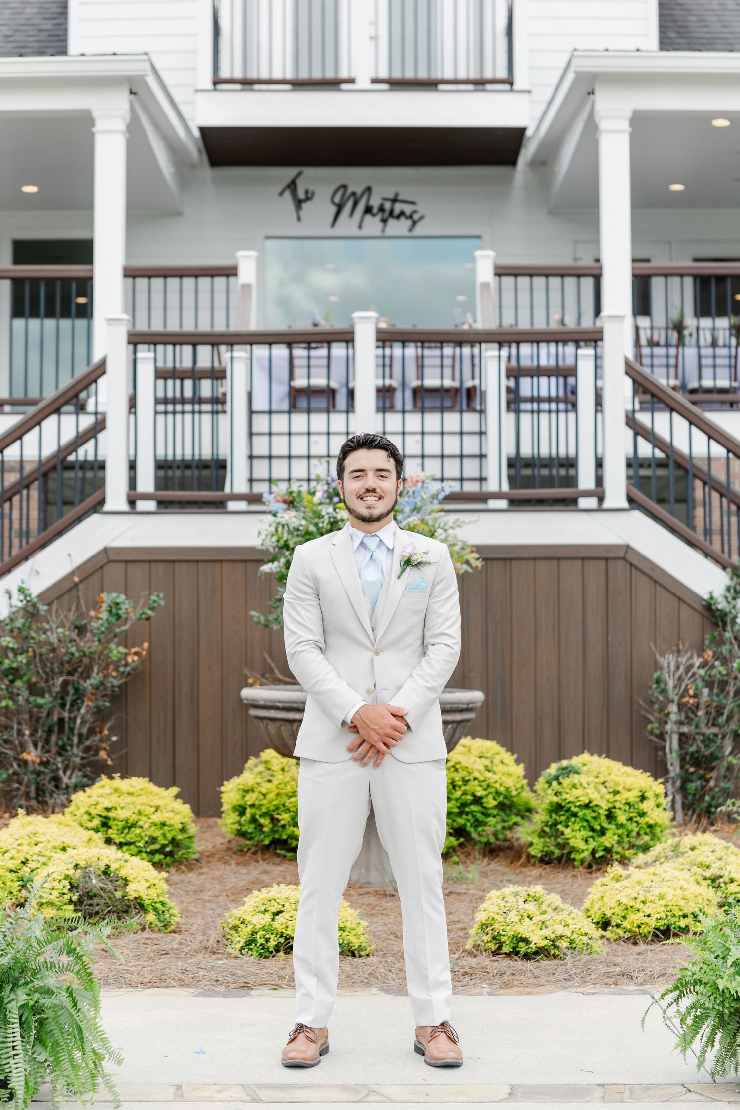 Groom in light gray suit with blue tie poses confidently outside elegant white wedding venue building.