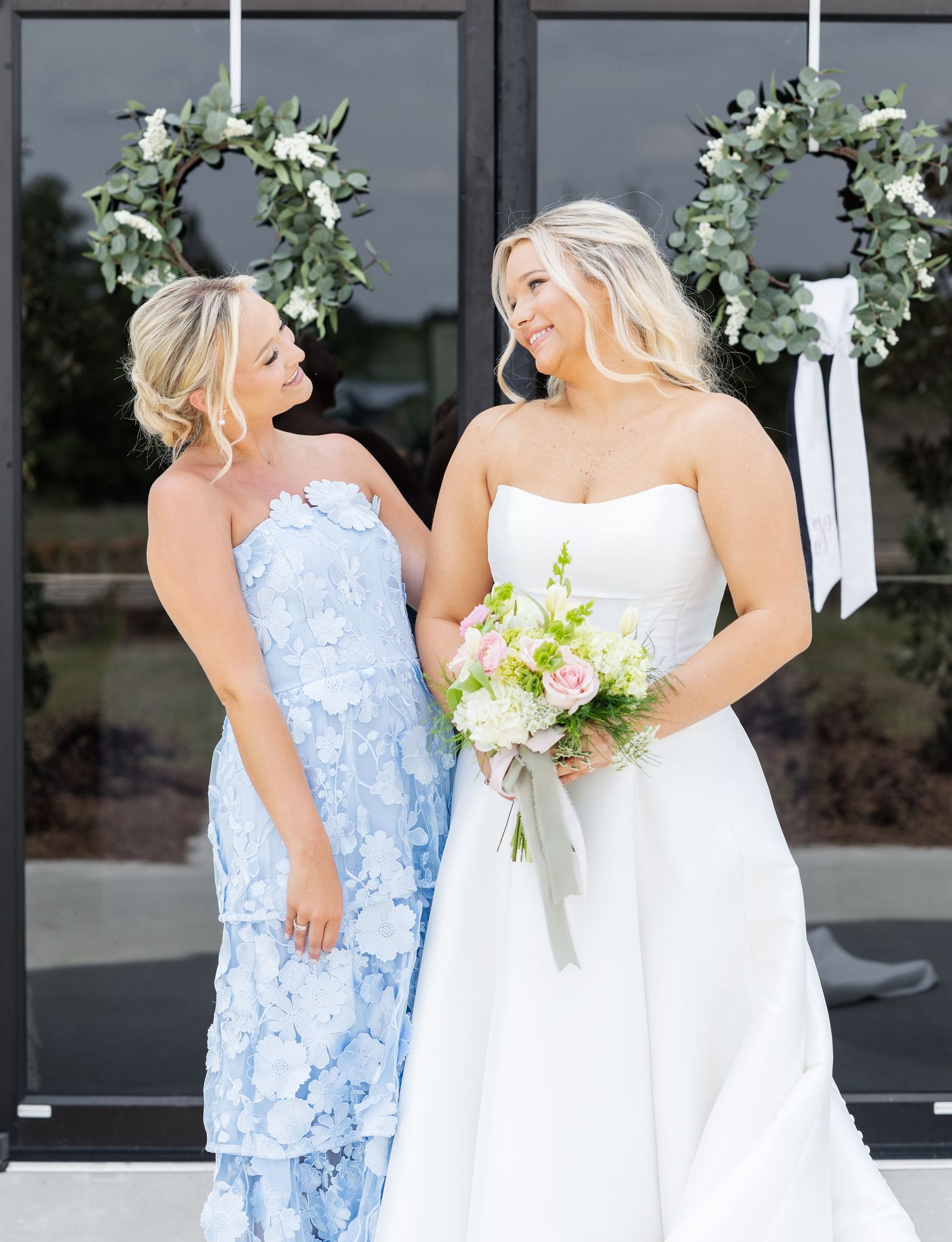 Bride in strapless white gown and woman in blue floral dress smile together before wedding ceremony.