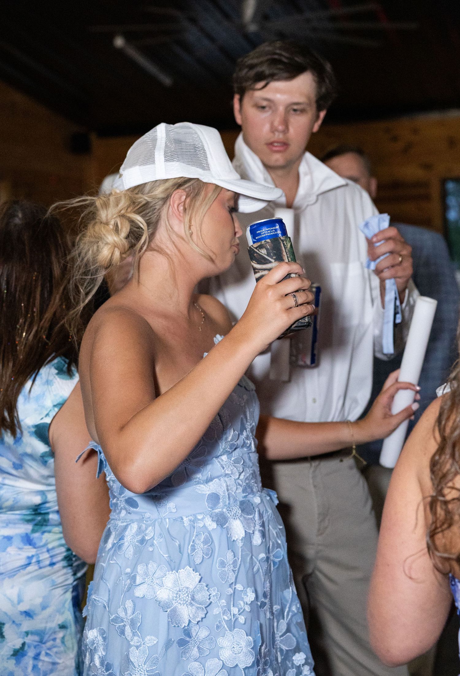 Young woman in blue floral dress and white cap drinking at a wedding reception celebration.