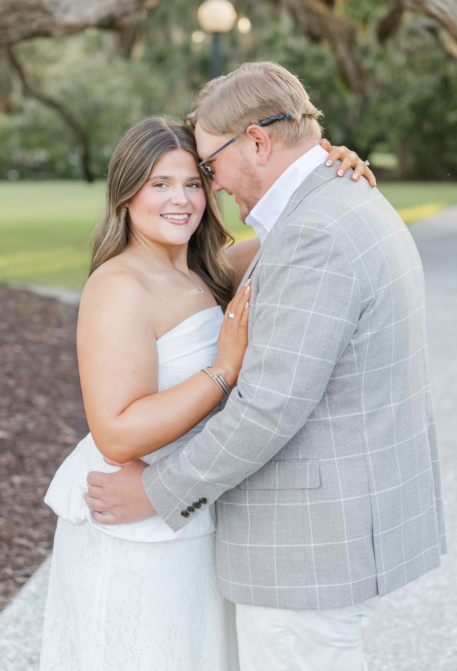 A young woman in a white strapless dress embraces an older man in a gray plaid blazer outdoors.