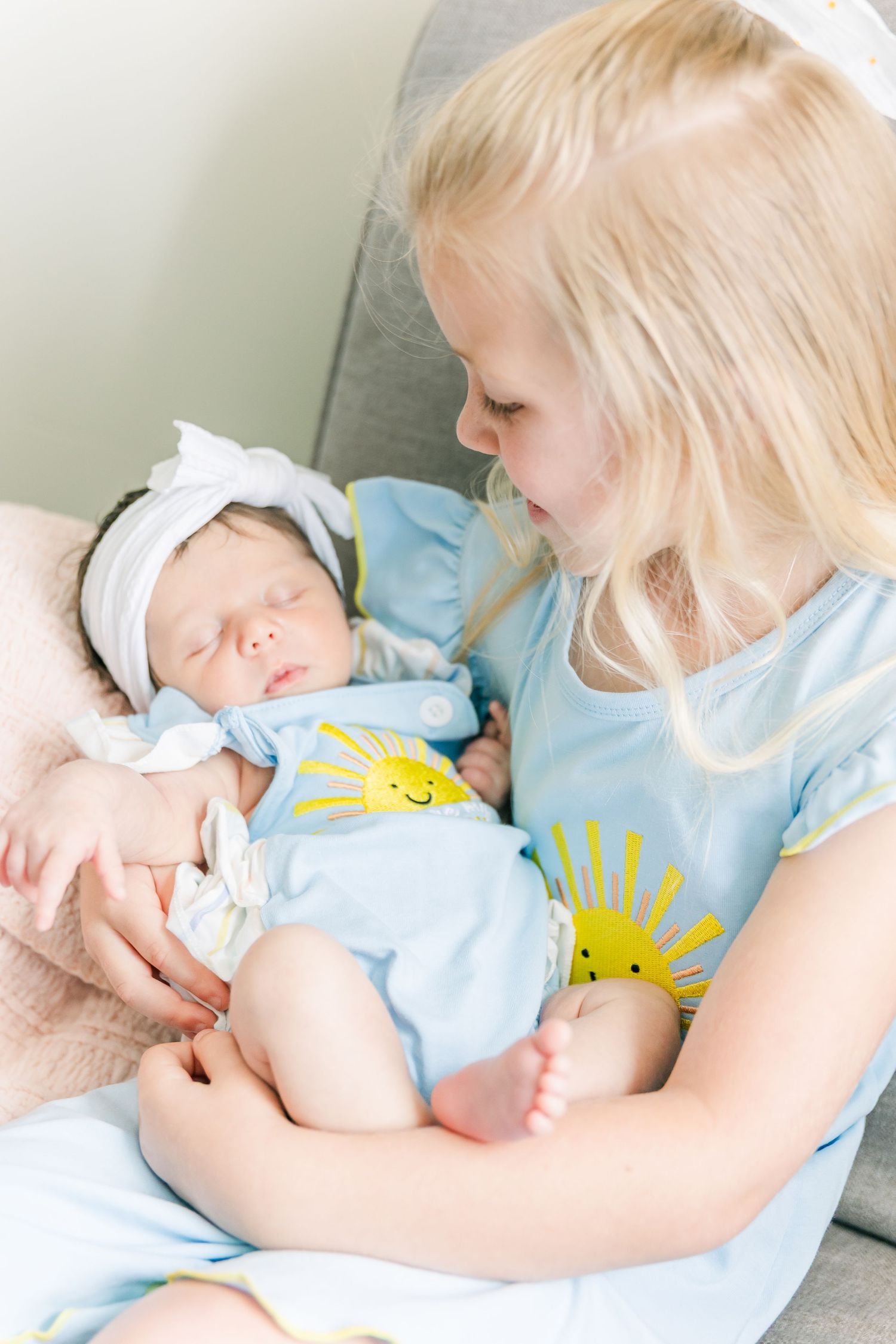 Young girl in blue shirt tenderly holds sleeping newborn sibling wearing white headband.