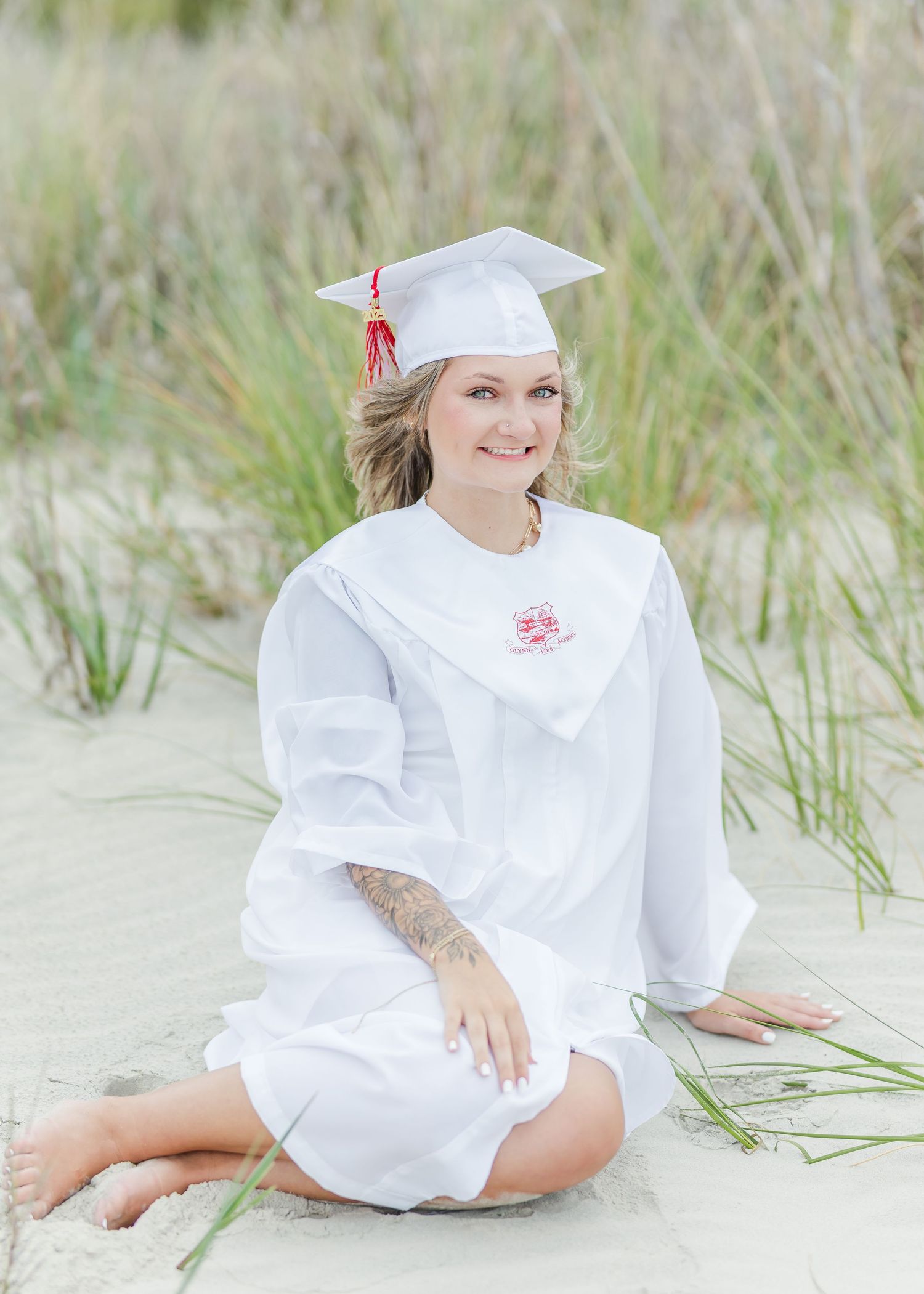 Graduate in white cap and gown sits on beach sand surrounded by sea grass.