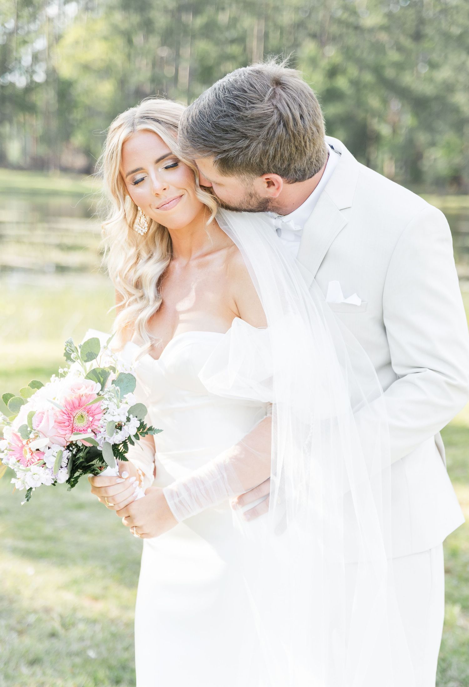Romantic outdoor wedding photo with couple embracing in all-white attire and holding pink bouquet.