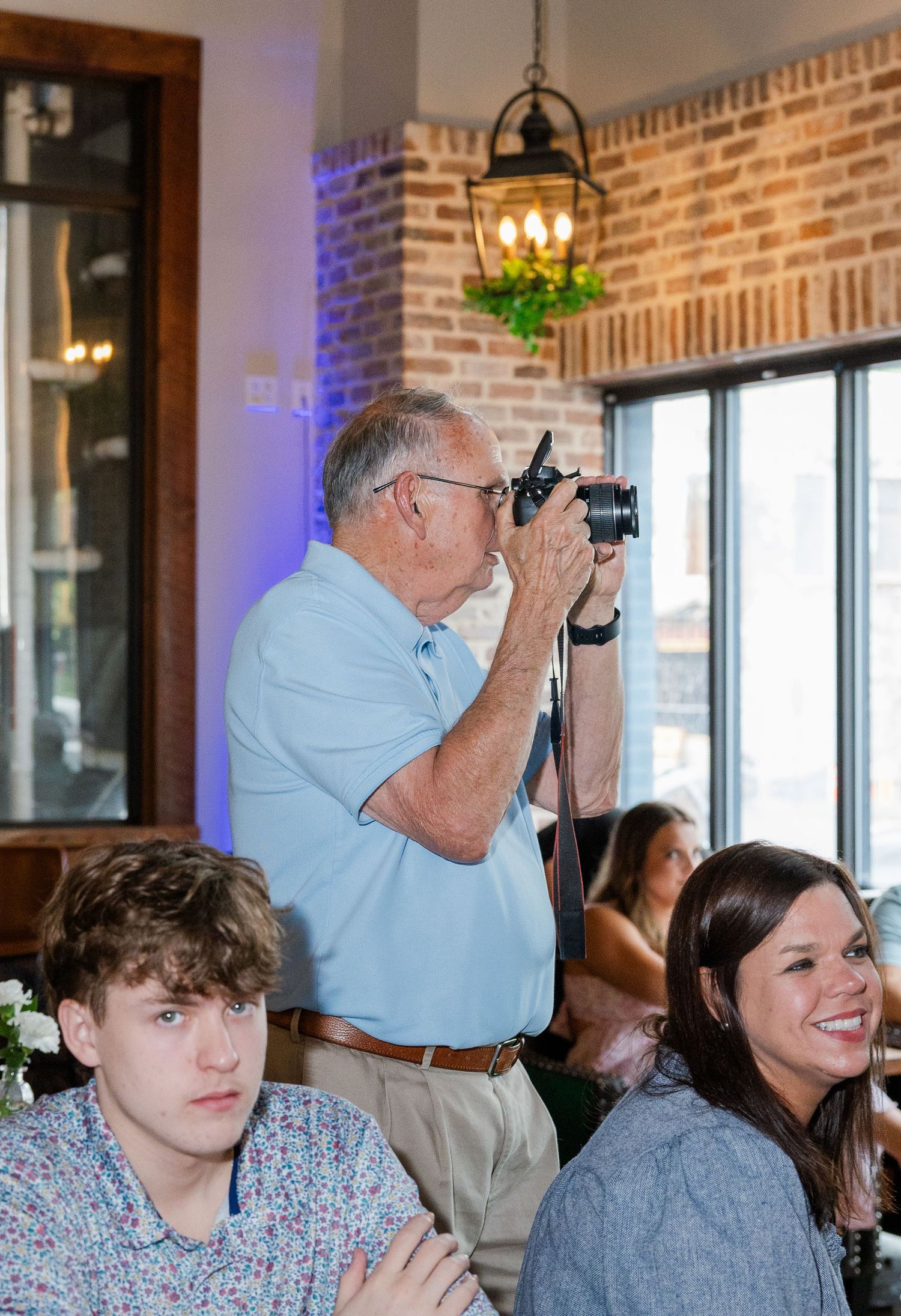 An elderly man in a light blue polo shirt photographs guests at an indoor event with brick walls and warm lighting.
