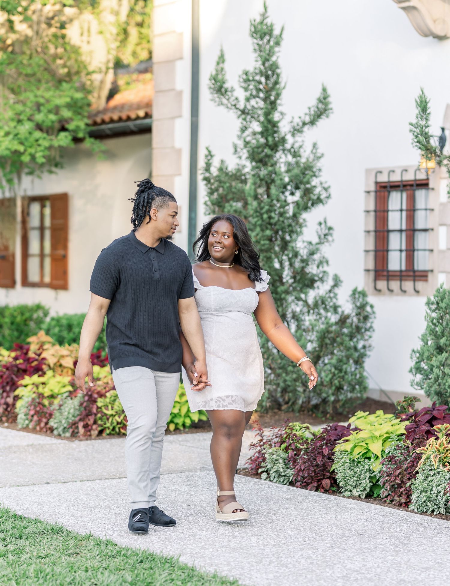 A couple holds hands and walks along a garden path, smiling at each other, near a white building with lush greenery.