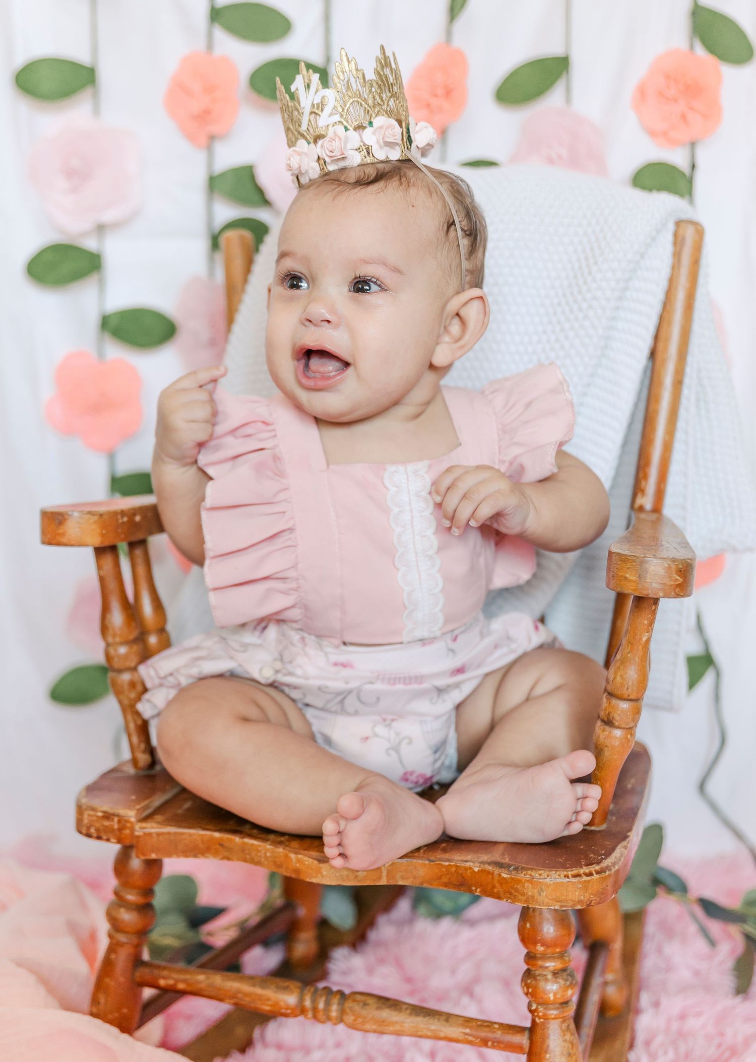 Baby wearing gold crown and pink outfit smiles while sitting in vintage wooden rocking chair.