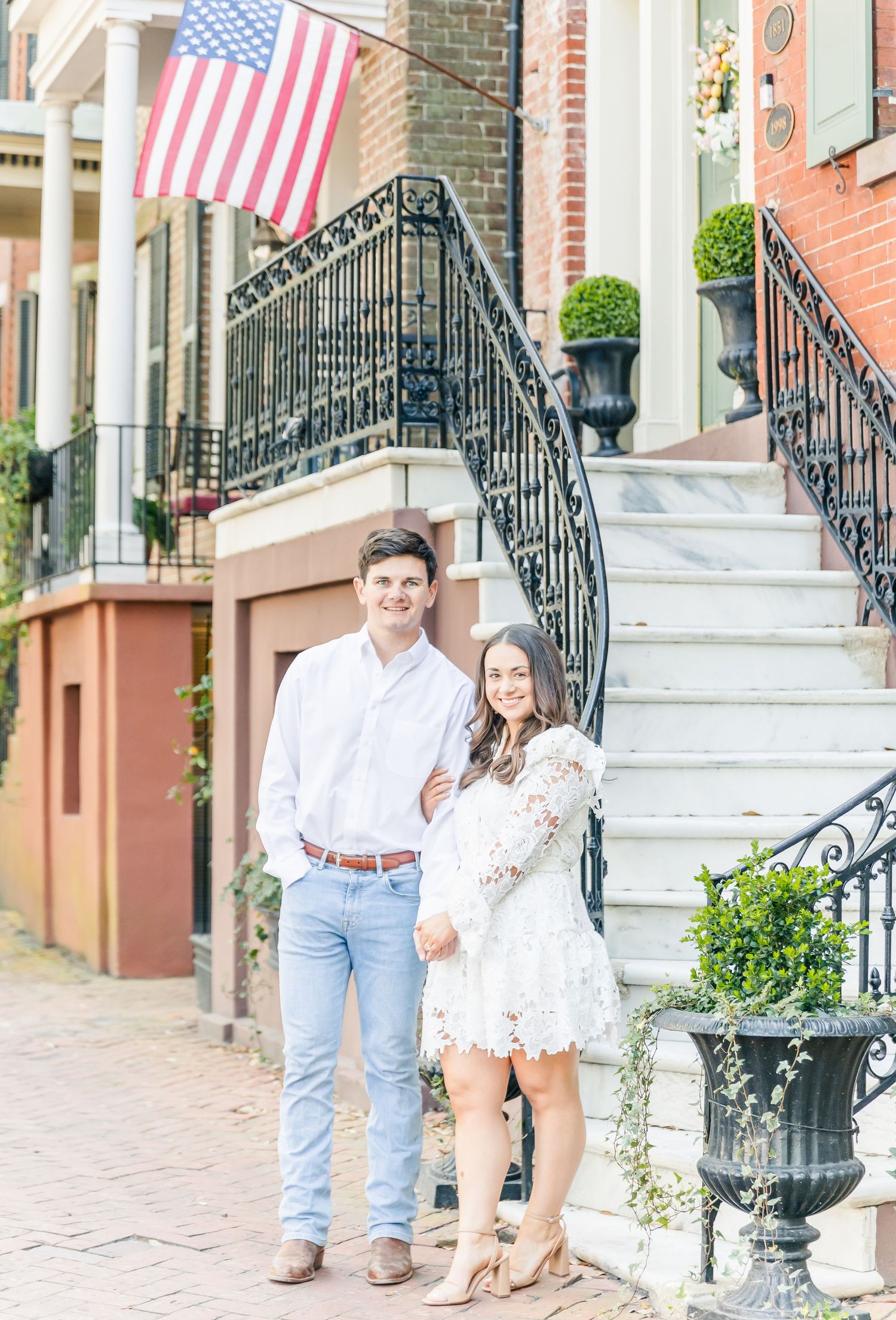 A couple stands on historic brick steps with ornate iron railings and an American flag in Charleston, South Carolina.