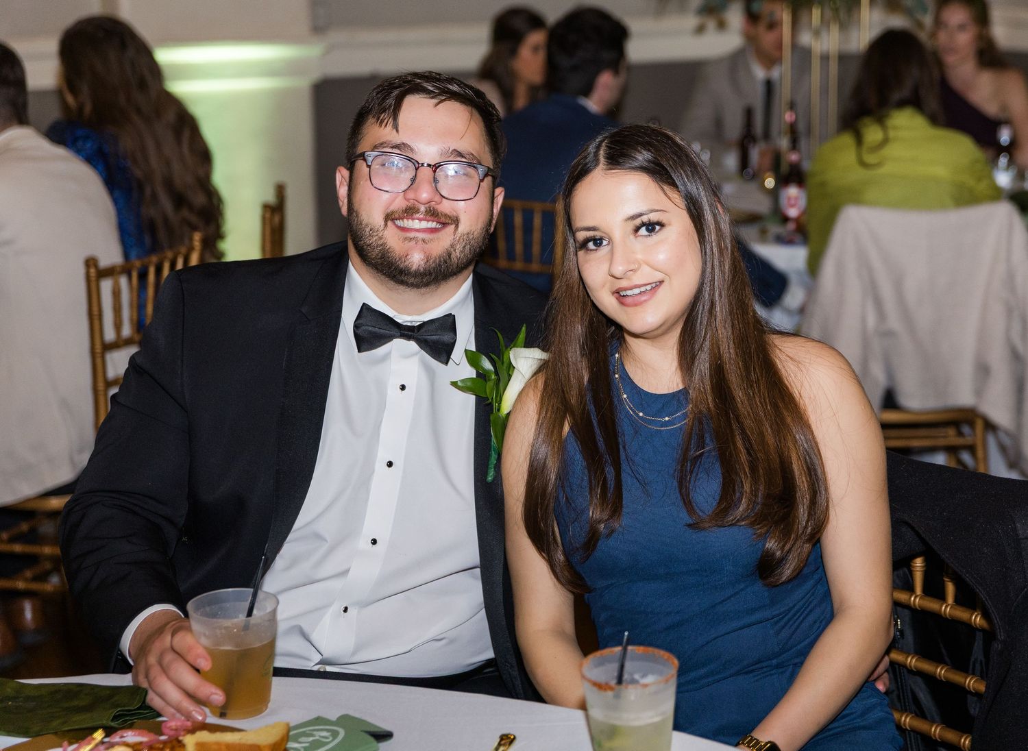Man in tuxedo and woman in blue dress smiling at wedding reception table.