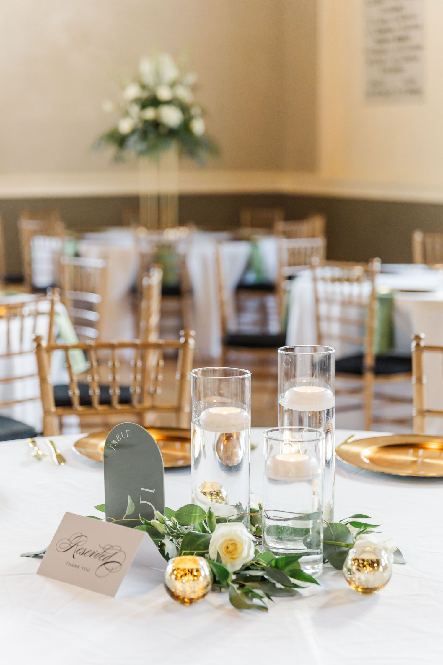 Elegant floating candle centerpiece with white roses and greenery garland on white reception table with gold chargers.