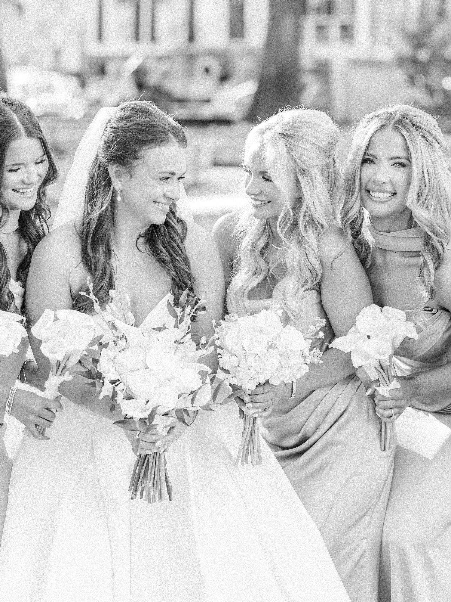 Black and white photo of a bride and three bridesmaids in satin gowns laughing together while holding white floral bouquets.