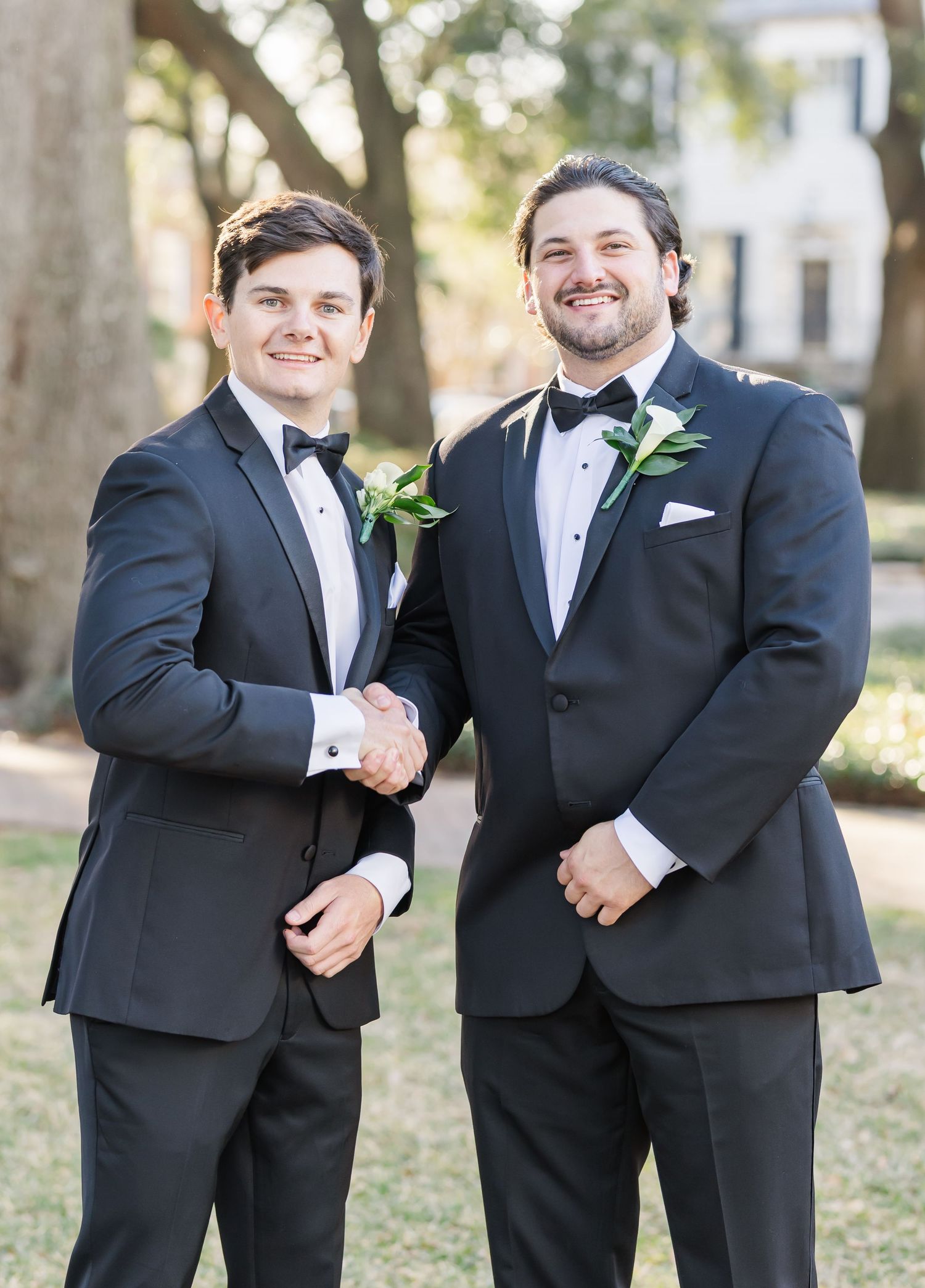 Two groomsmen in black tuxedos with boutonnieres shake hands and smile at outdoor wedding.