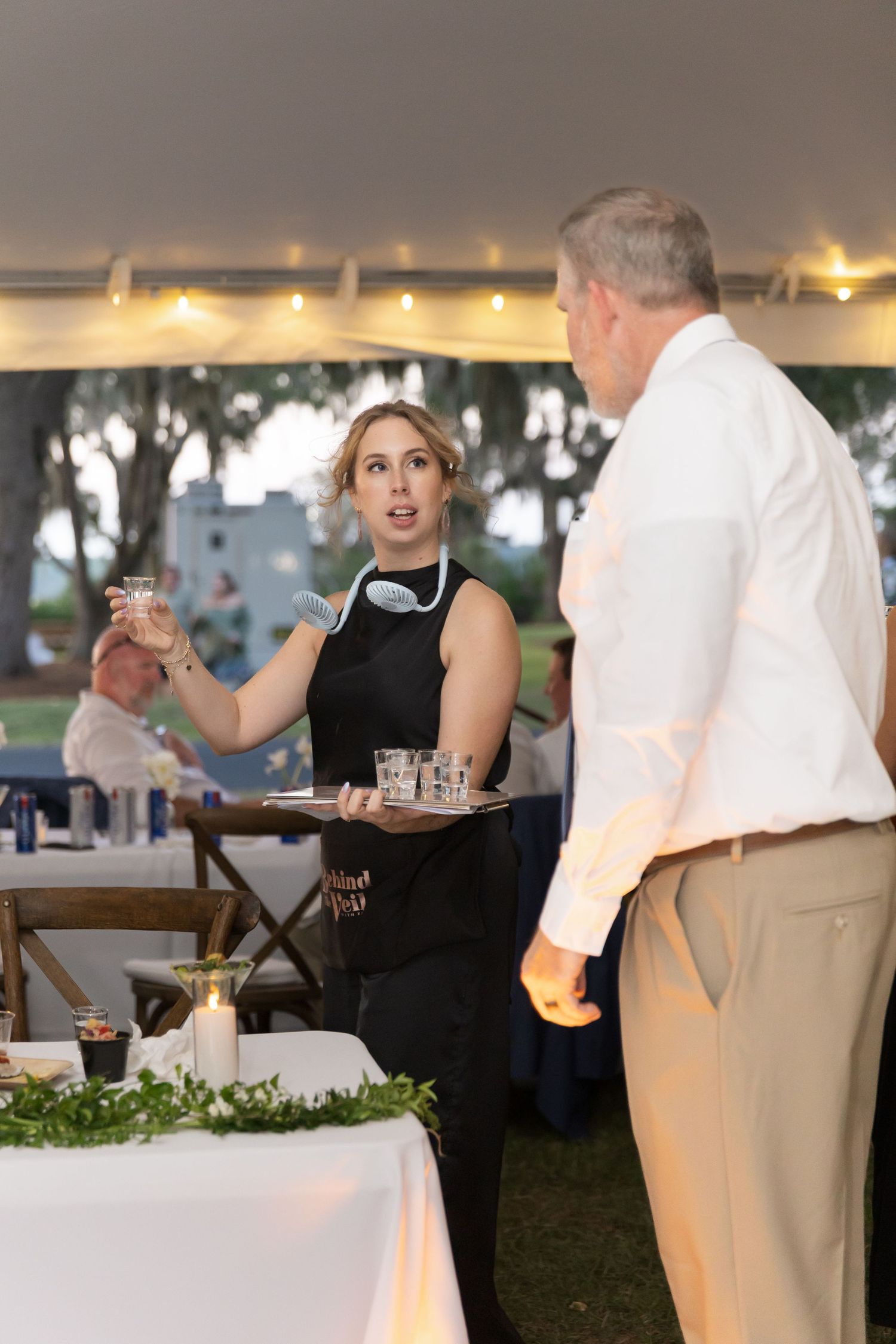 A server in a black dress carries a tray of drinks during an outdoor evening event with string lights overhead.
