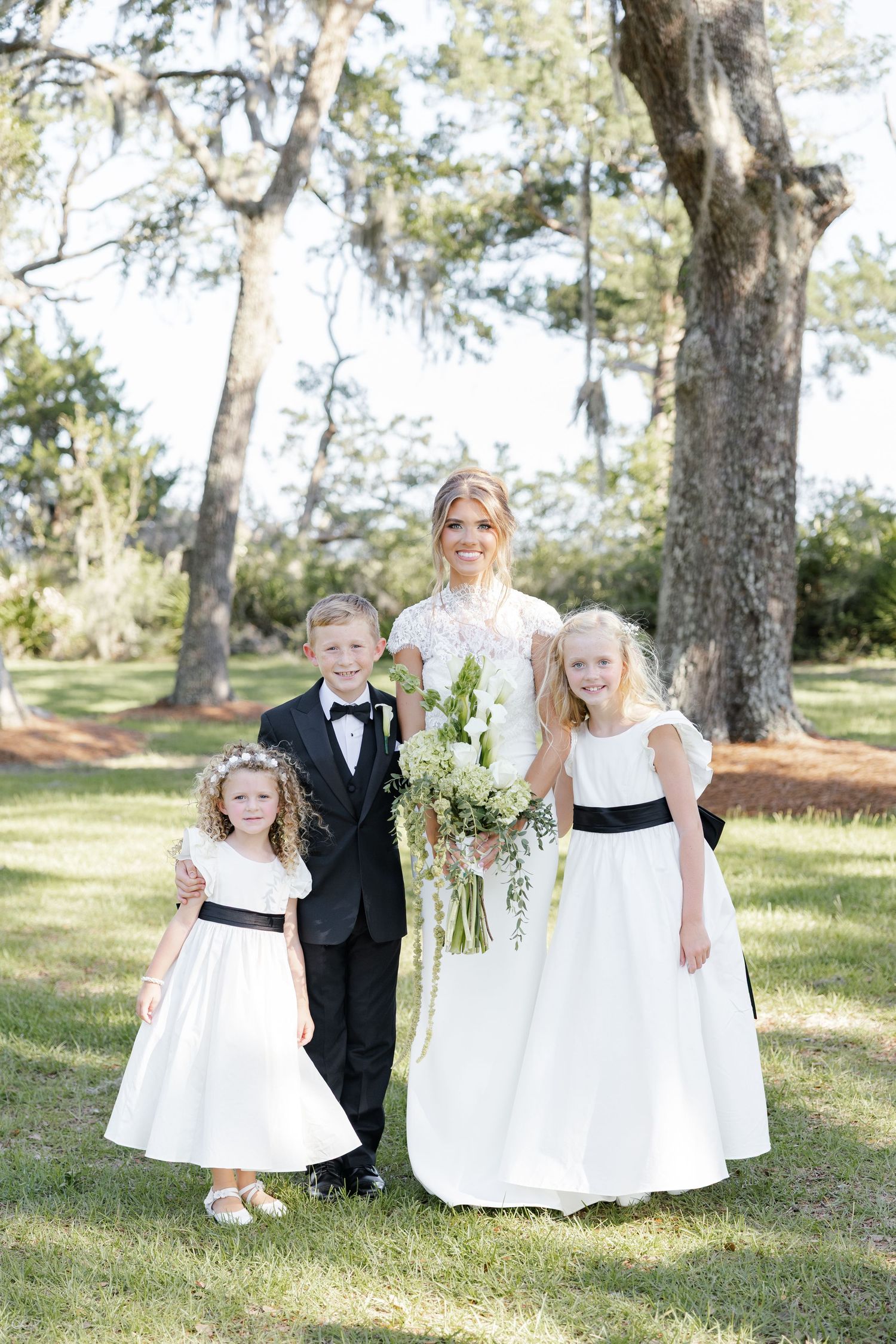 Bride in lace gown holds bouquet with flower girls in white dresses and ring bearer in tuxedo outdoors.