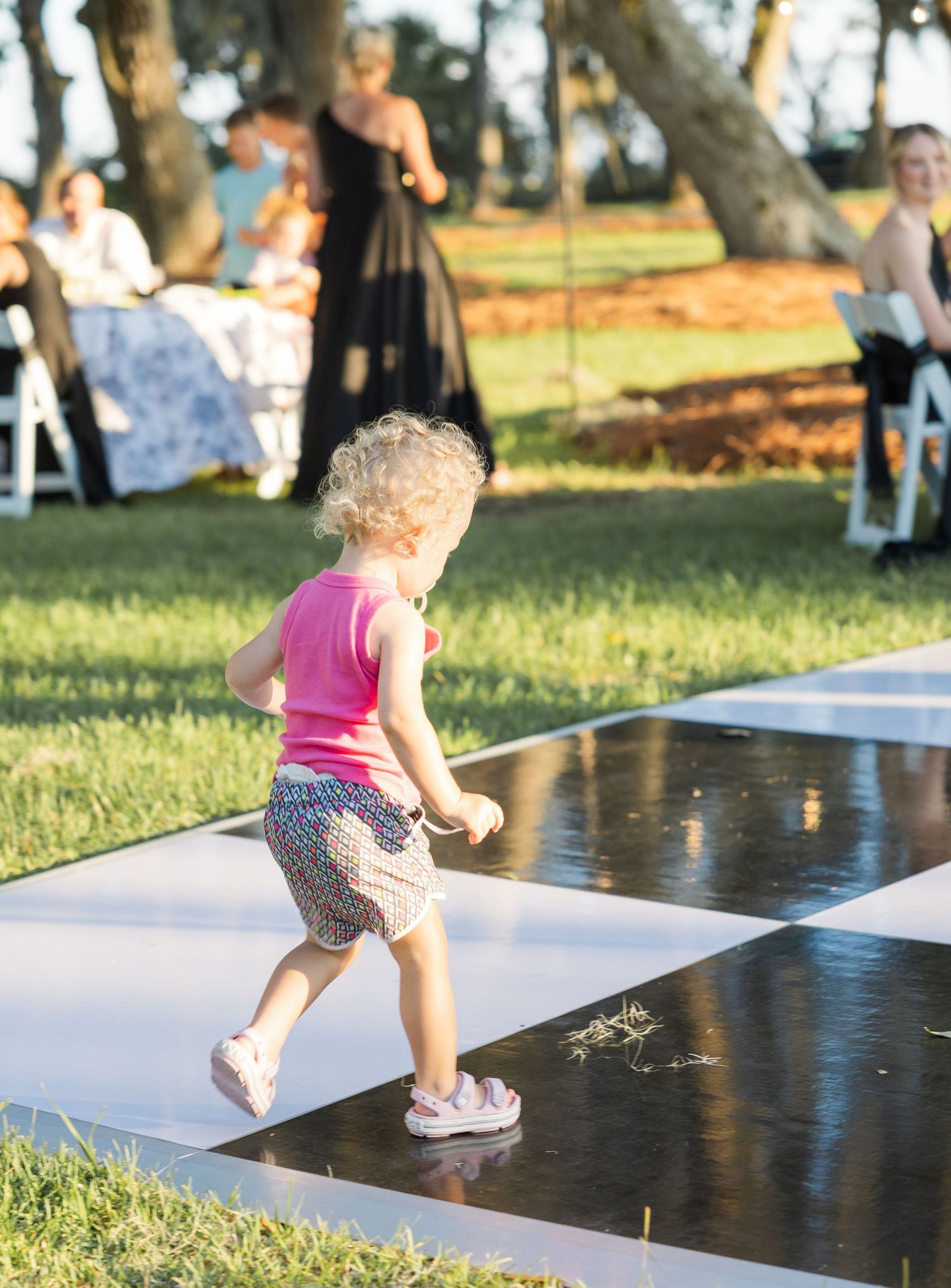 Toddler girl in pink top dancing on checkered dance floor at outdoor wedding reception.
