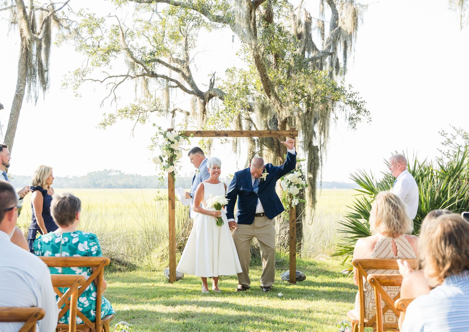 Guests seated in wooden chairs at an outdoor wedding ceremony under Spanish moss draped trees by the waterfront.