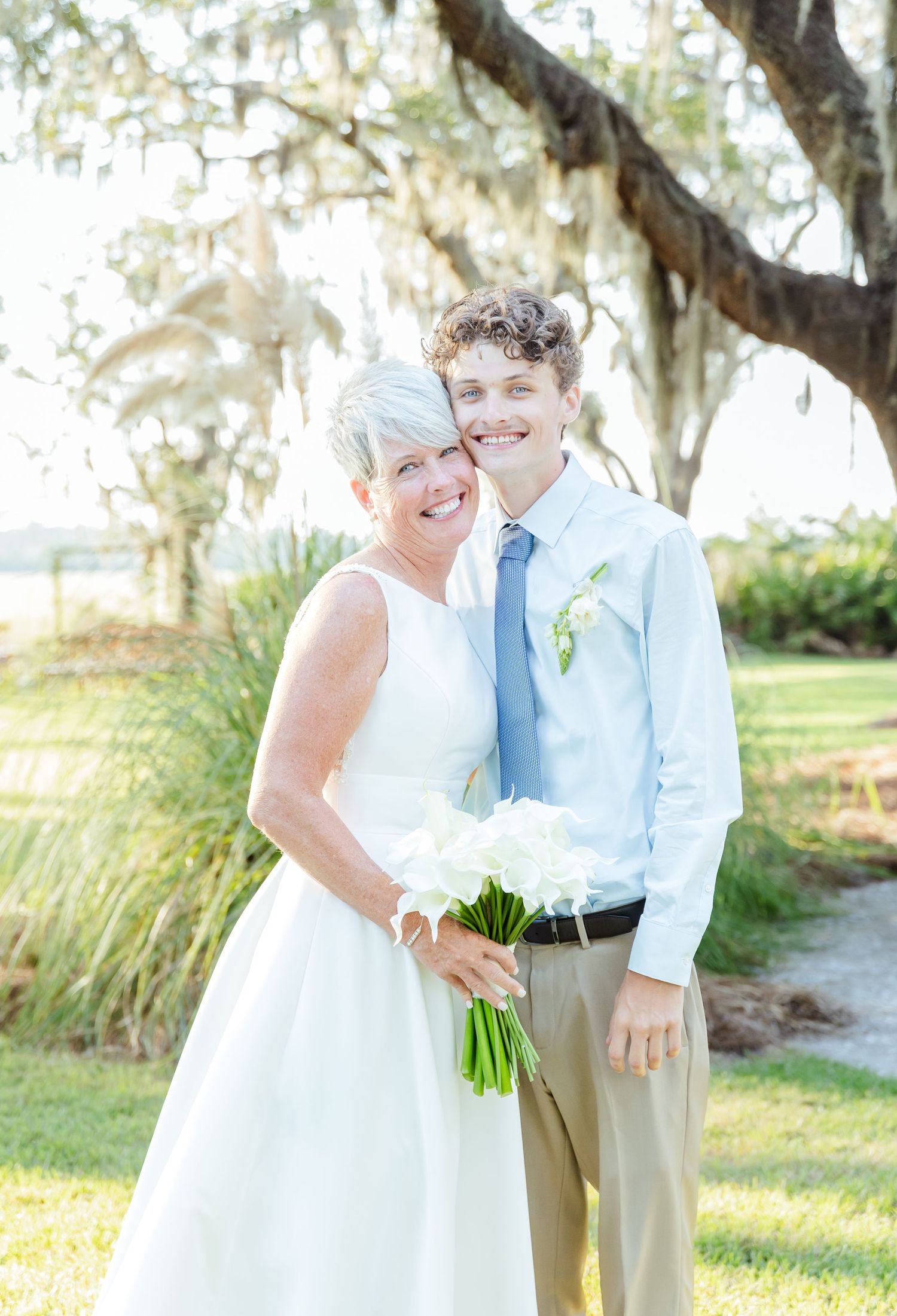 A couple in wedding attire smiles together under Spanish moss draped trees in a romantic outdoor setting.