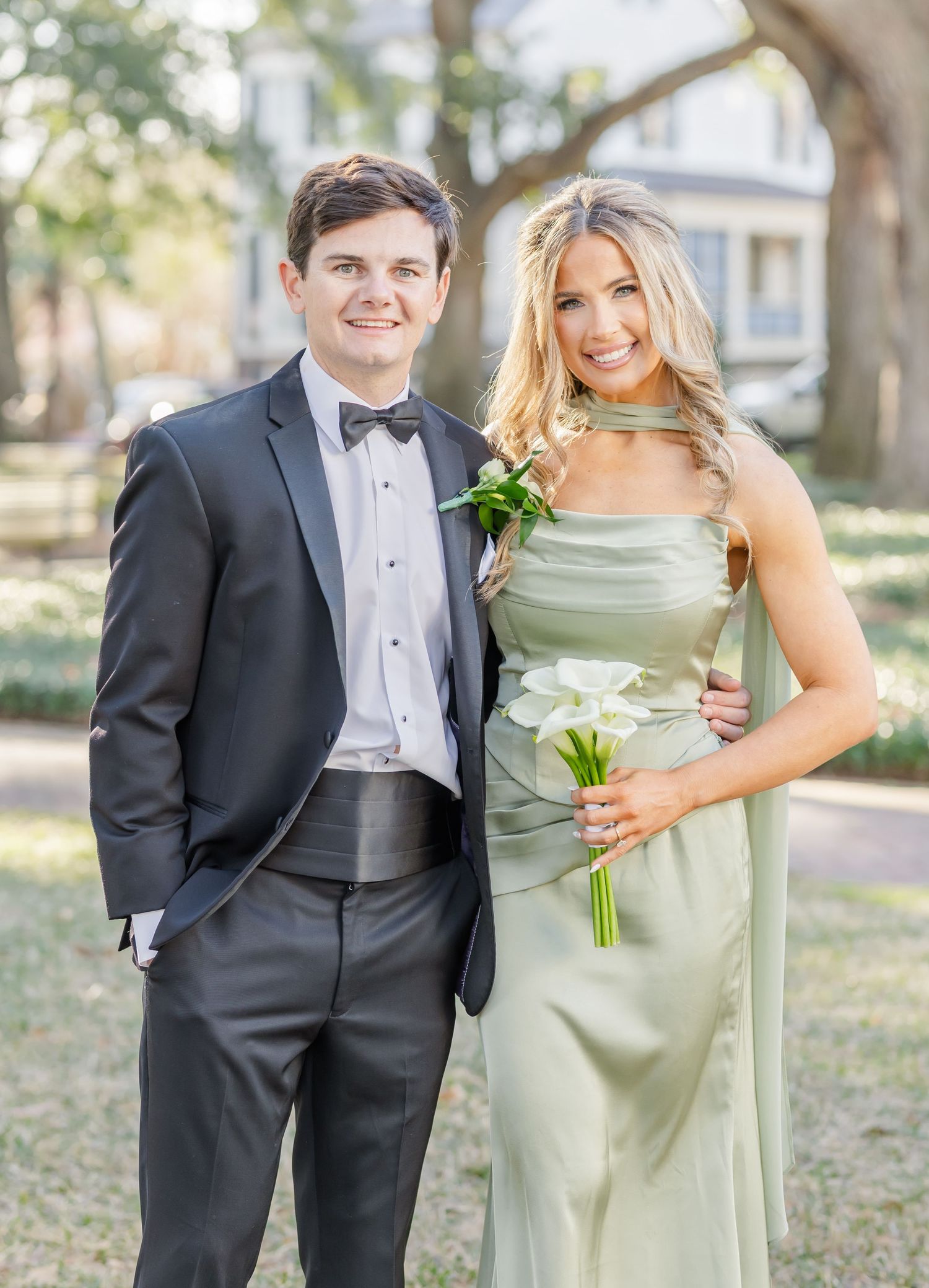 Wedding party members in formal attire pose together under trees at an outdoor venue.