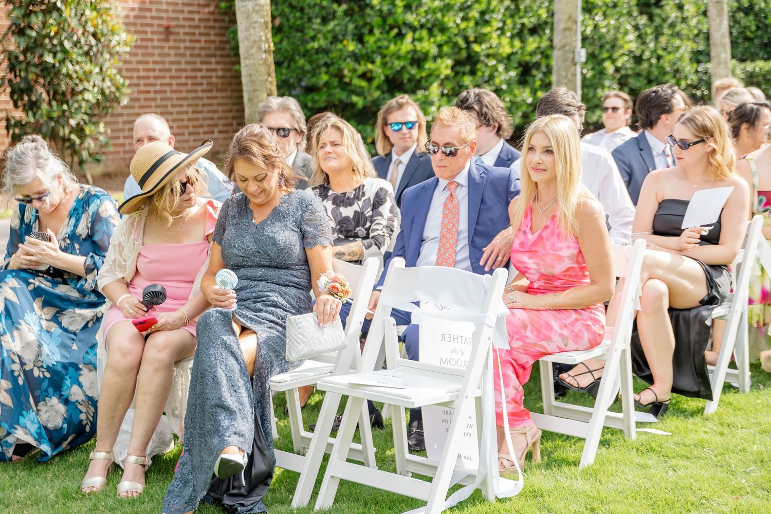 Wedding guests seated in white folding chairs on a lawn watch an outdoor ceremony on a sunny summer day.