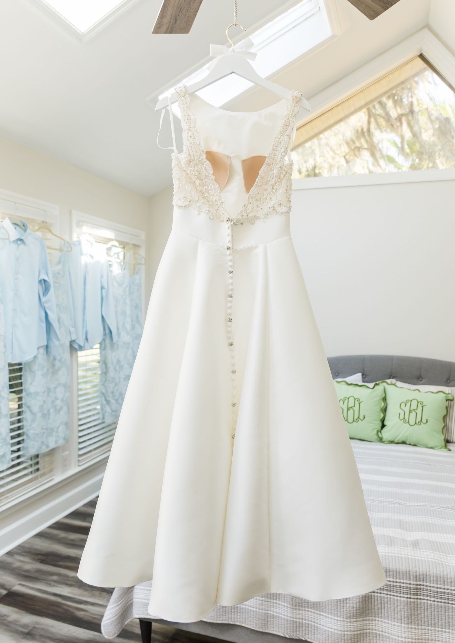 Multiple white christening or baptism dresses displayed on hangers against a window with sheer curtains.