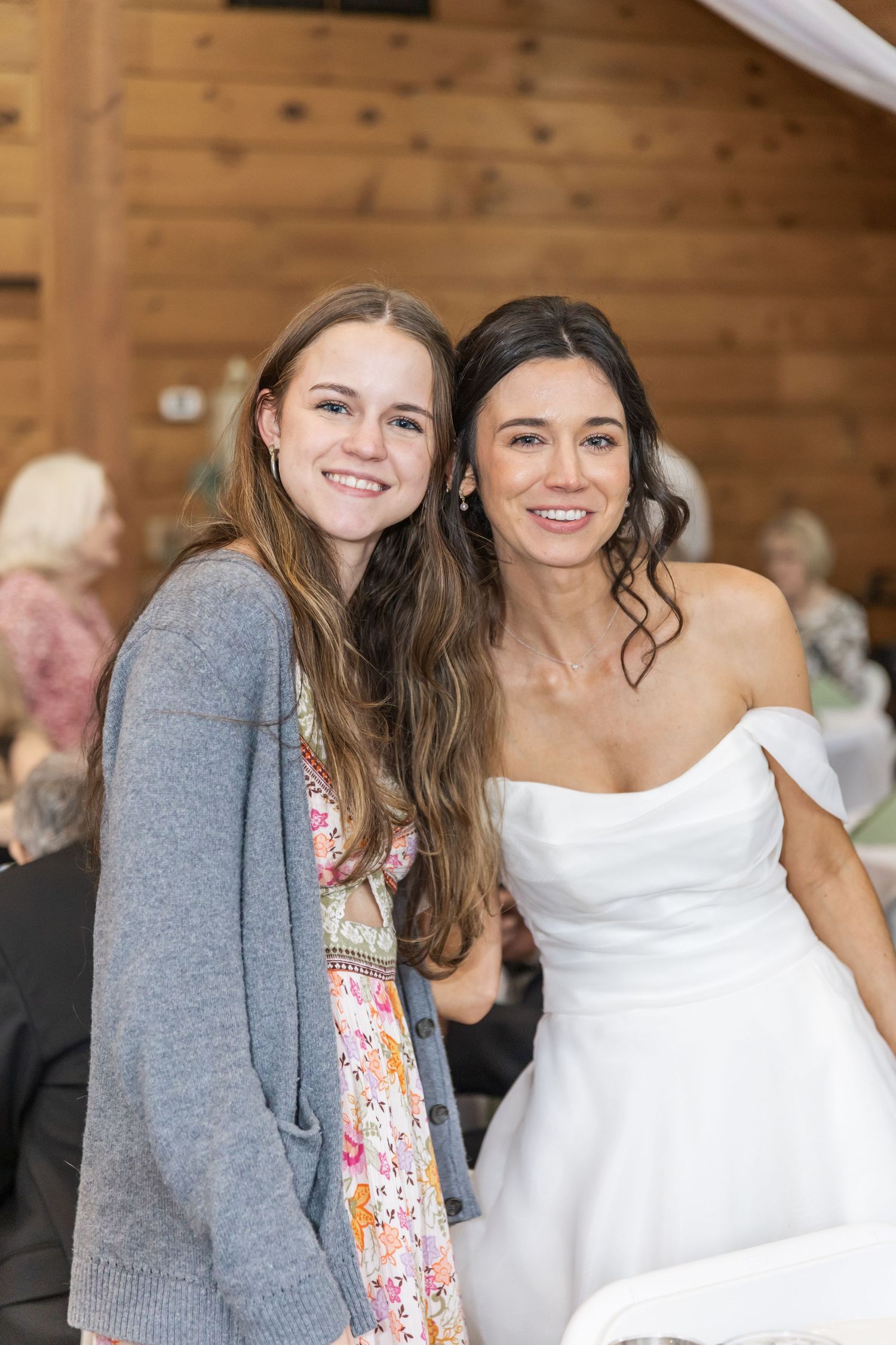 Two guests at a rustic wedding reception pose together, one wearing a floral dress and grey cardigan.