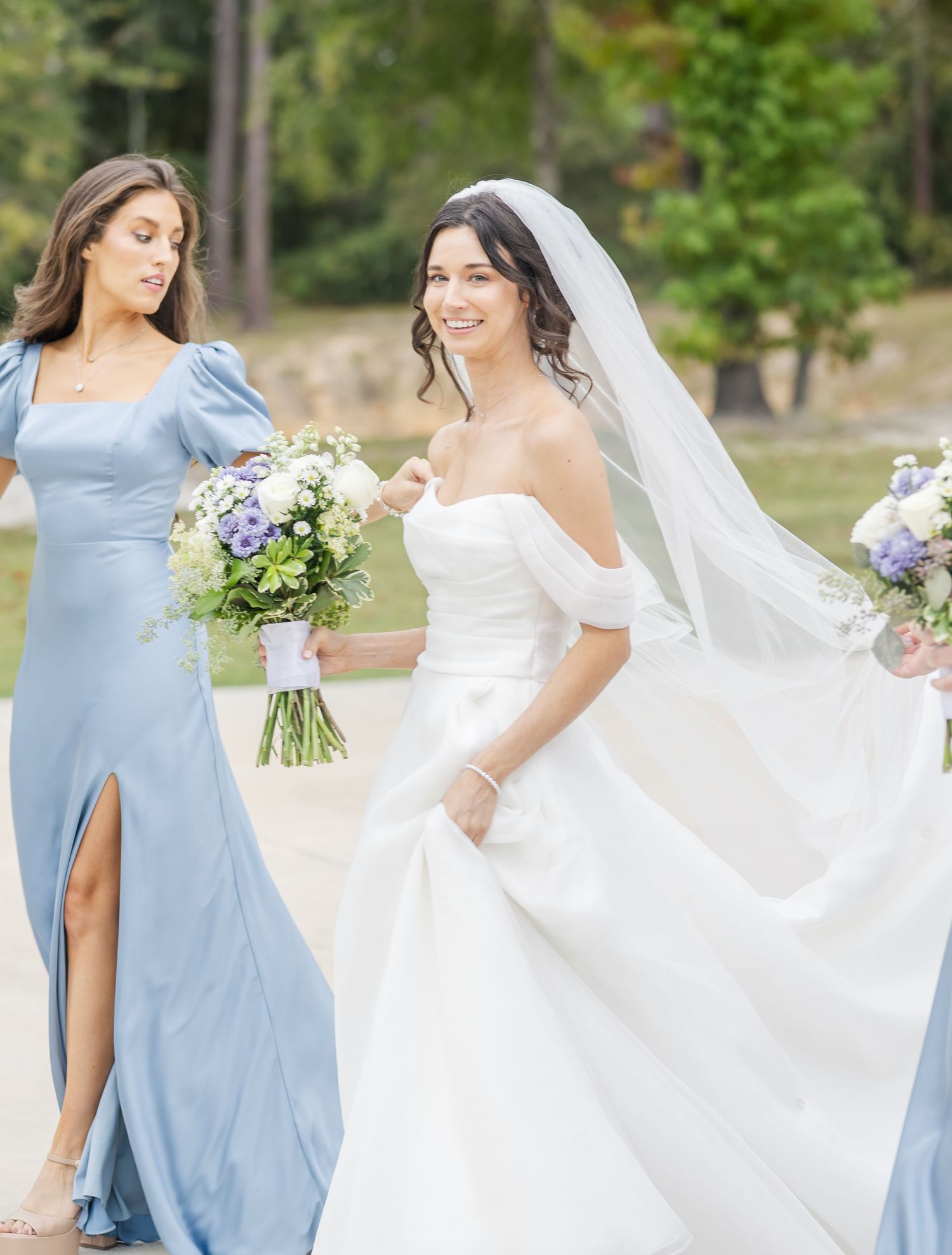 A bridal party in light blue bridesmaid dresses and white wedding gown pose together outdoors in a garden setting.
