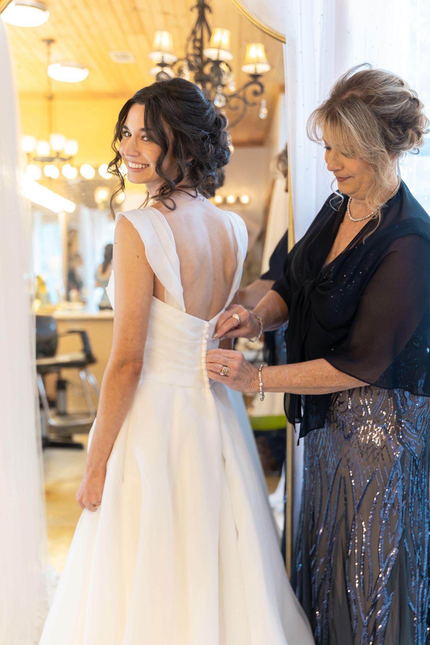 A bride in a white wedding dress gets help with final adjustments from a woman wearing a dark sequined gown.