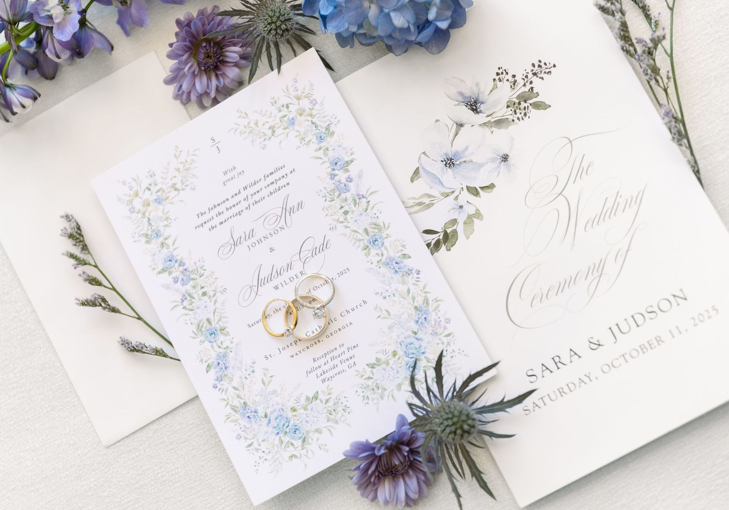 Wedding invitation suite with delicate blue flowers and calligraphy laid out on a white surface with dried florals.