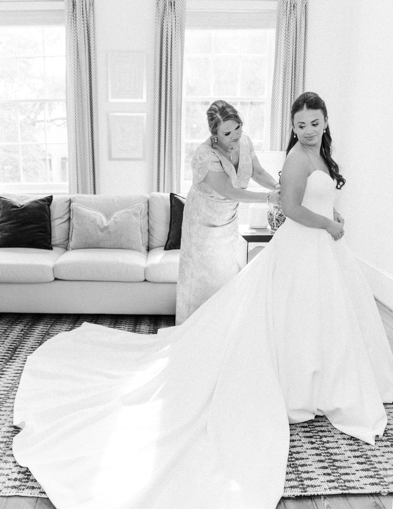 A black and white photo showing a wedding dress fitting moment in a bright room with a modern couch and patterned rug.