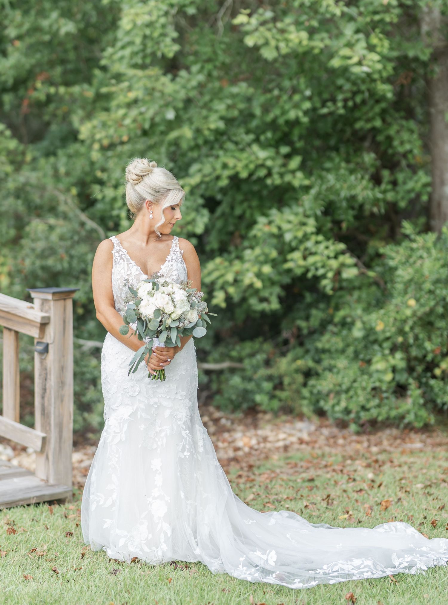 A bride in a white lace wedding gown poses gracefully in a garden setting with flowing train and bouquet.