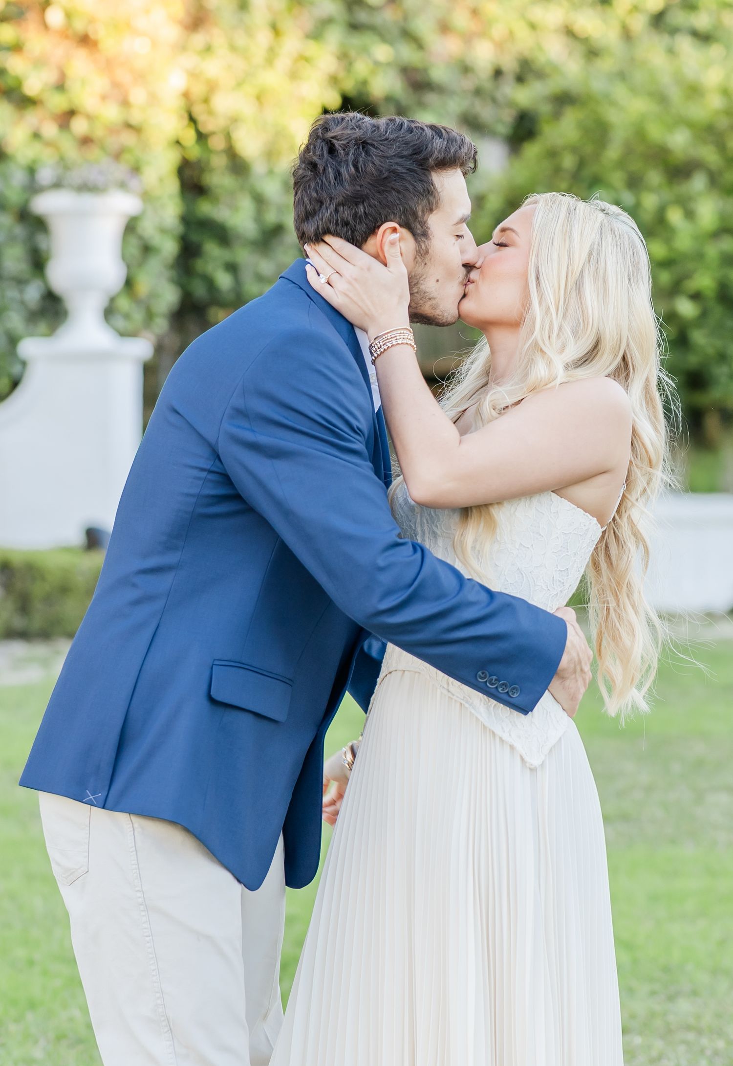 A couple in formal wedding attire share romantic kisses in a garden setting with white decorative elements.