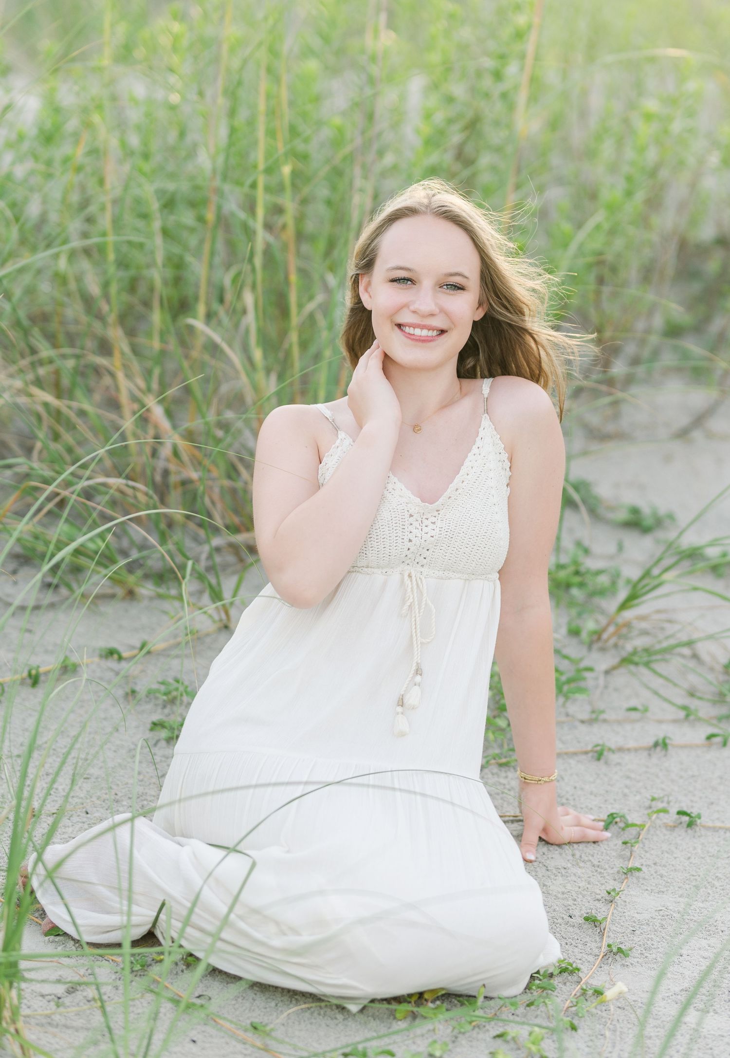 A person in a white sundress poses on sandy dunes with beach grass in the background.
