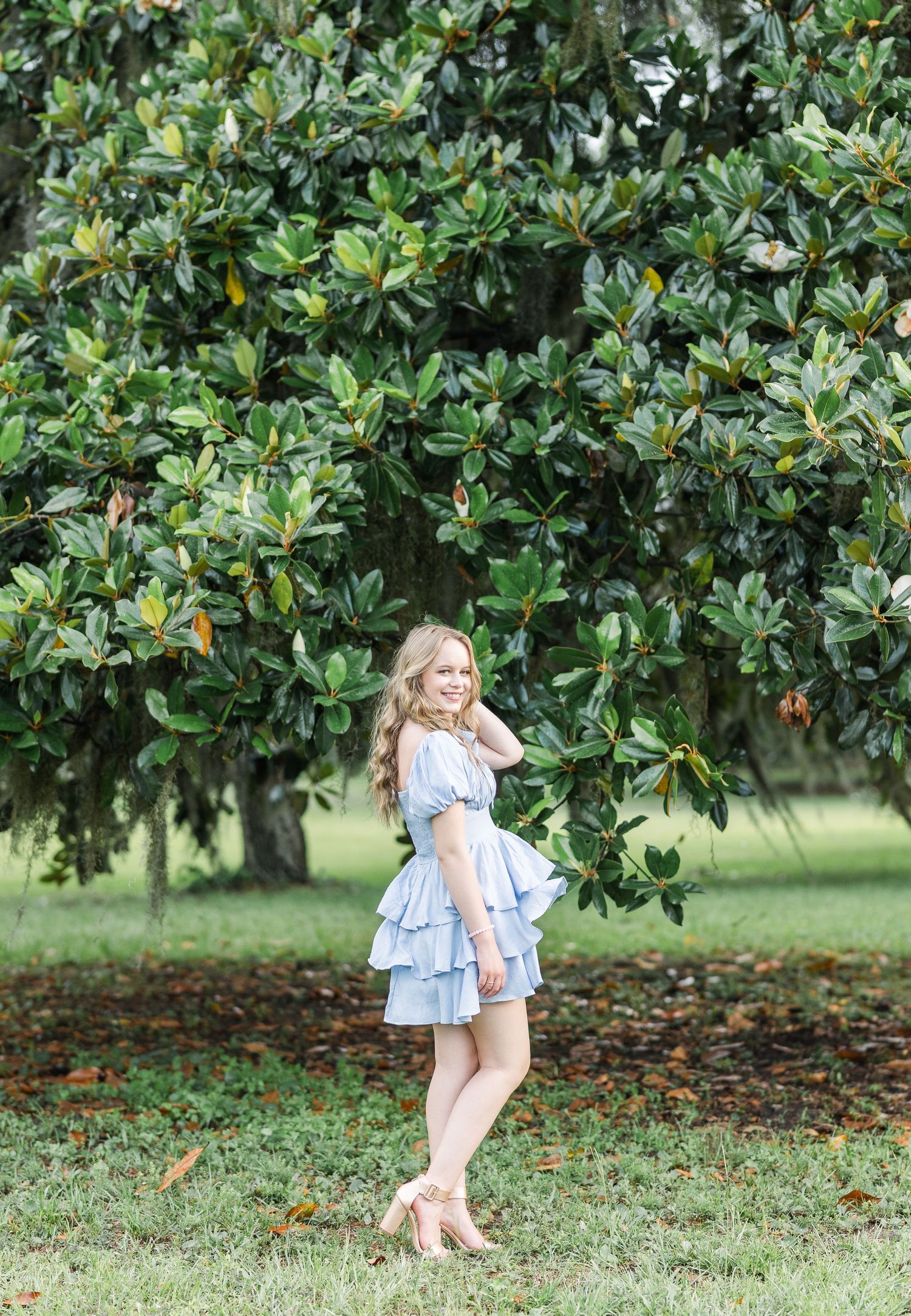A woman in a light blue ruffled dress poses gracefully beneath a lush magnolia tree in a garden setting.