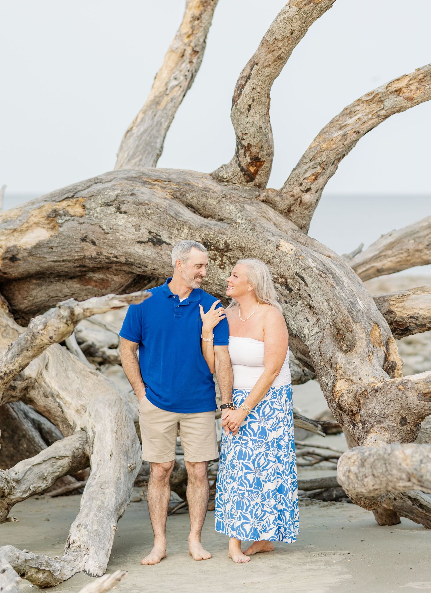 A couple stands together on a beach by large driftwood logs, wearing blue and white summer attire.