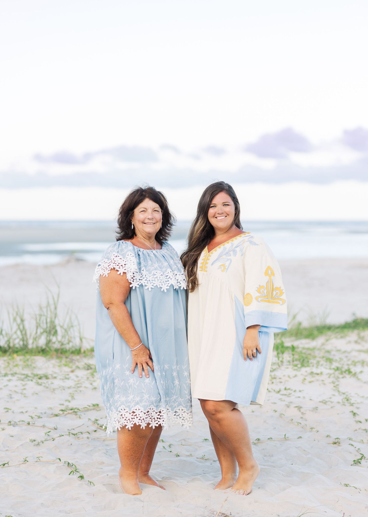 Two people in soft blue and white summer dresses pose together on a sandy beach at dusk.